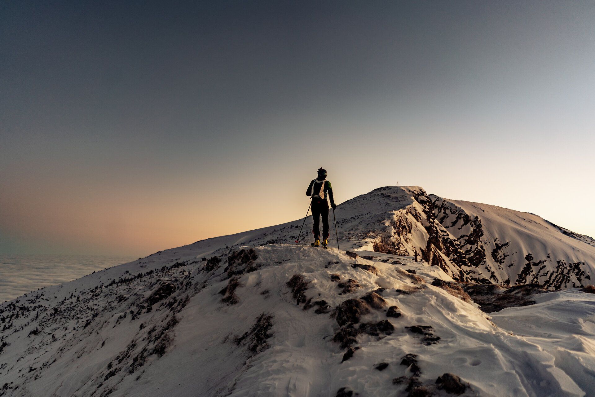Skitourengeher bei Sonnenaufgang auf einem verschneiten Berggrat; im Hintergrund weite Winter-Berglandschaft über einem Meer aus Wolken.