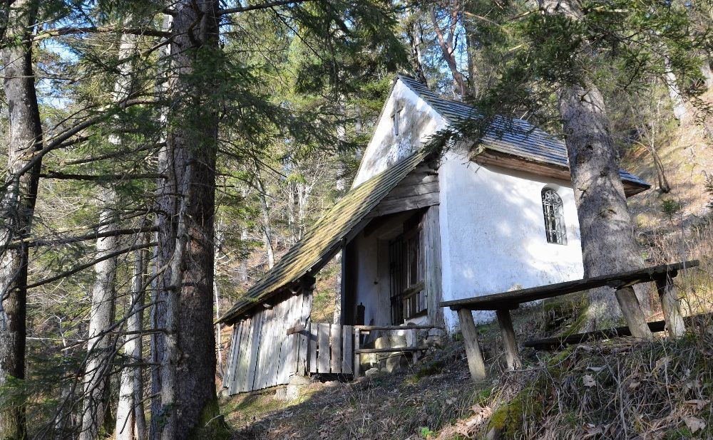 Eine kleine weiße Kapelle im Wald, umgeben von Bäumen und einem Holztisch im Vordergrund.