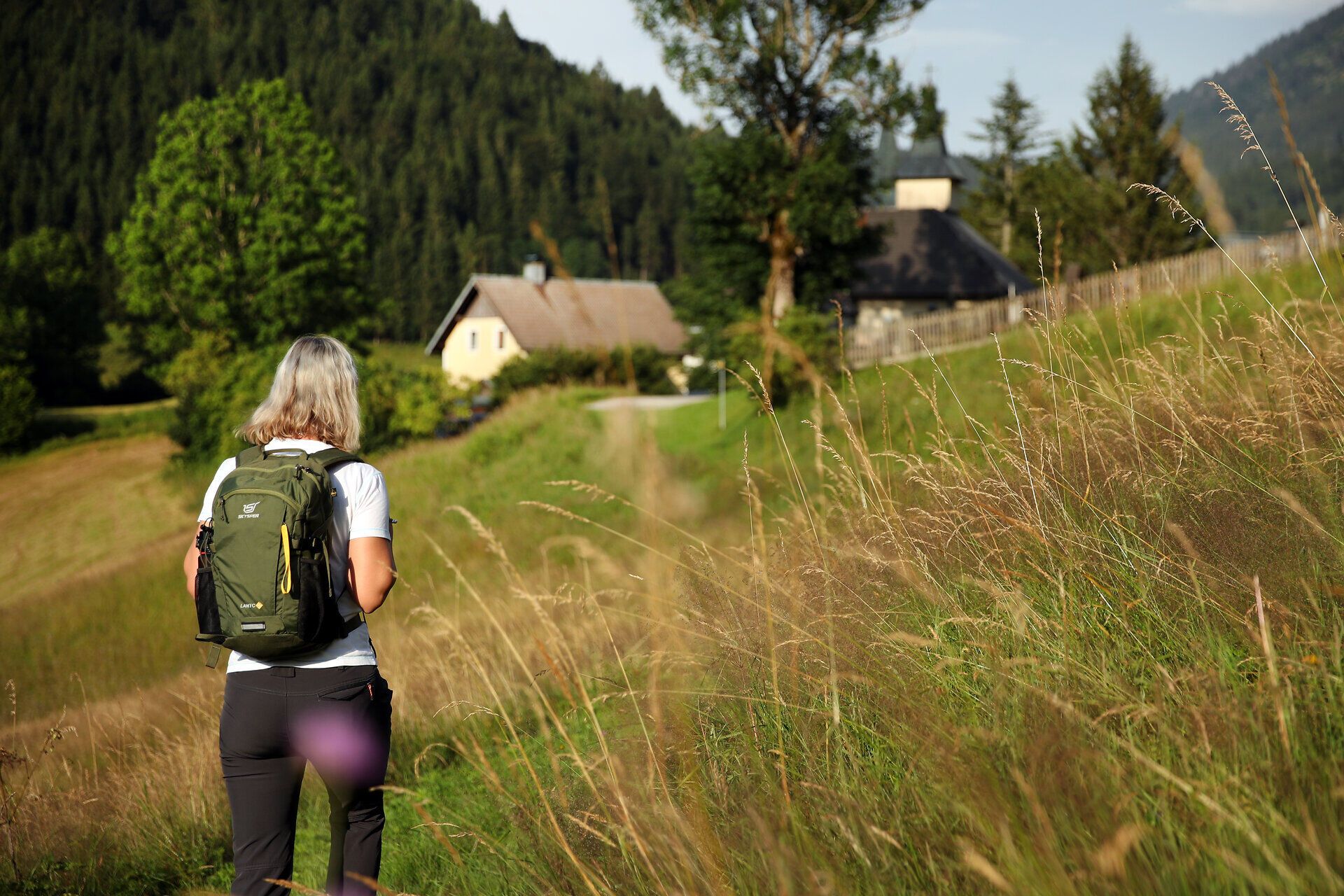 Pilgerin auf steilem Wiesenhang mit Blick auf Bauernhof.