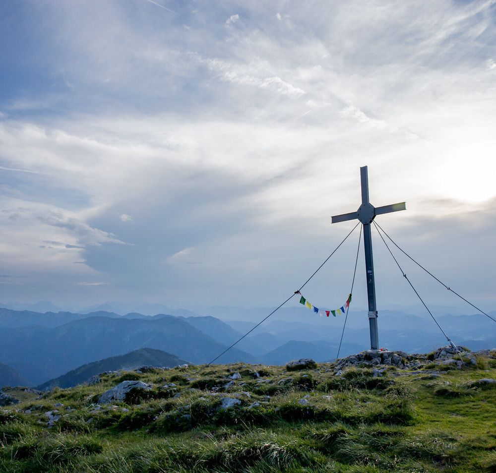 Ein majestätisches Gipfelkreuz thront über den sanften Hügeln, umgeben von einer atemberaubenden Berglandschaft. Die Wolken ziehen sanft über den Himmel und verleihen der Szenerie eine friedliche Atmosphäre, während die frische Bergluft zum Wandern einlädt.