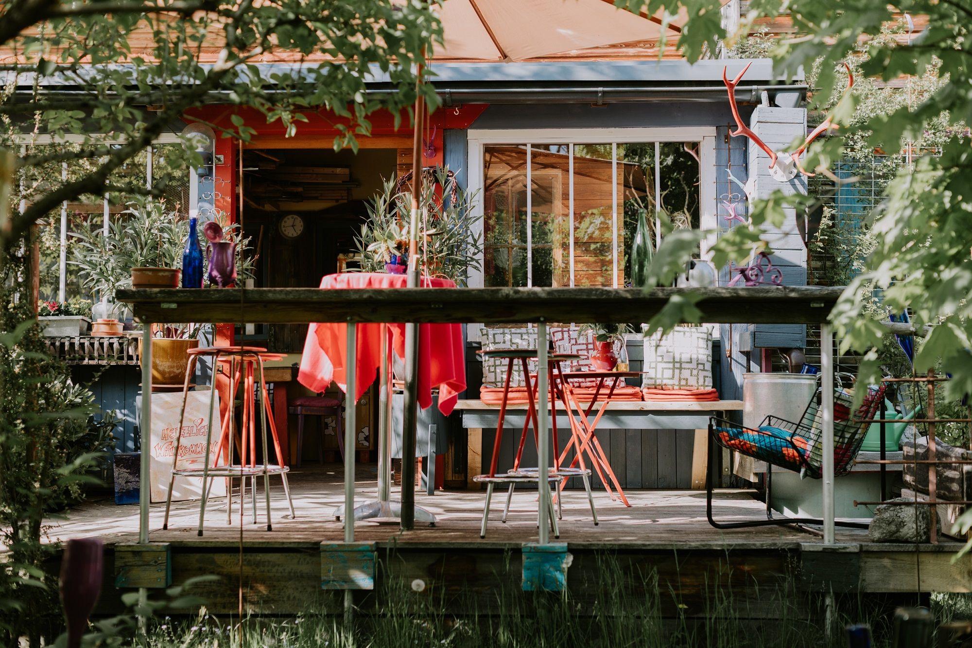 Bunte Terrasse vor der Location mit roten Stühlen und Tisch, umgeben von Pflanzen und Dekorationen.