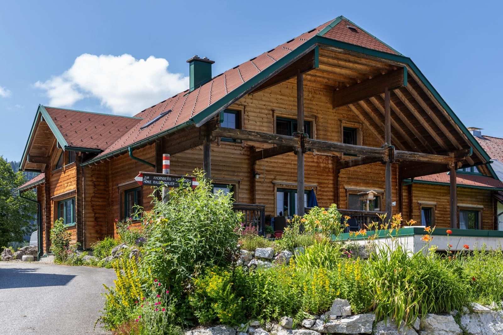 Ein traditionelles Holzhaus mit einem roten Dach, umgeben von bunten Blumen und grünen Pflanzen, unter einem blauen Himmel.