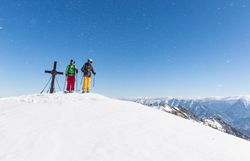 Auf dem schneebedeckten Gipfel genießen zwei Skifahrer die frische Bergluft und den atemberaubenden Ausblick auf die umliegenden Alpen. Die strahlende Sonne und der klare blaue Himmel schaffen eine perfekte Kulisse für ein unvergessliches Winterabenteuer.