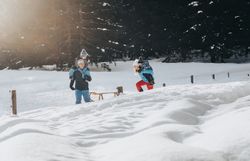 Eine Familie genießt den Winter in der verschneiten Landschaft der Ybbstaler Alpen. Die Kinder lachen und spielen im frischen Schnee, während die Sonne sanft durch die Bäume scheint und eine magische Atmosphäre schafft.