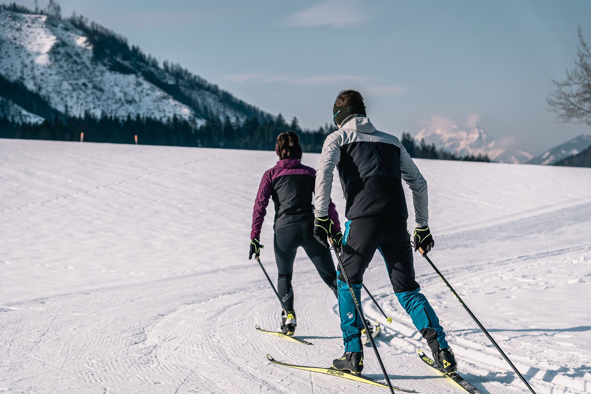 Die glitzernde Schneedecke erstreckt sich über die sanften Hügel der Ybbstaler Alpen, während zwei Skilangläufer in harmonischem Rhythmus durch die winterliche Landschaft gleiten. Die klare, kalte Luft und die majestätischen Berge im Hintergrund schaffen eine perfekte Kulisse für einen aktiven Winterurlaub.