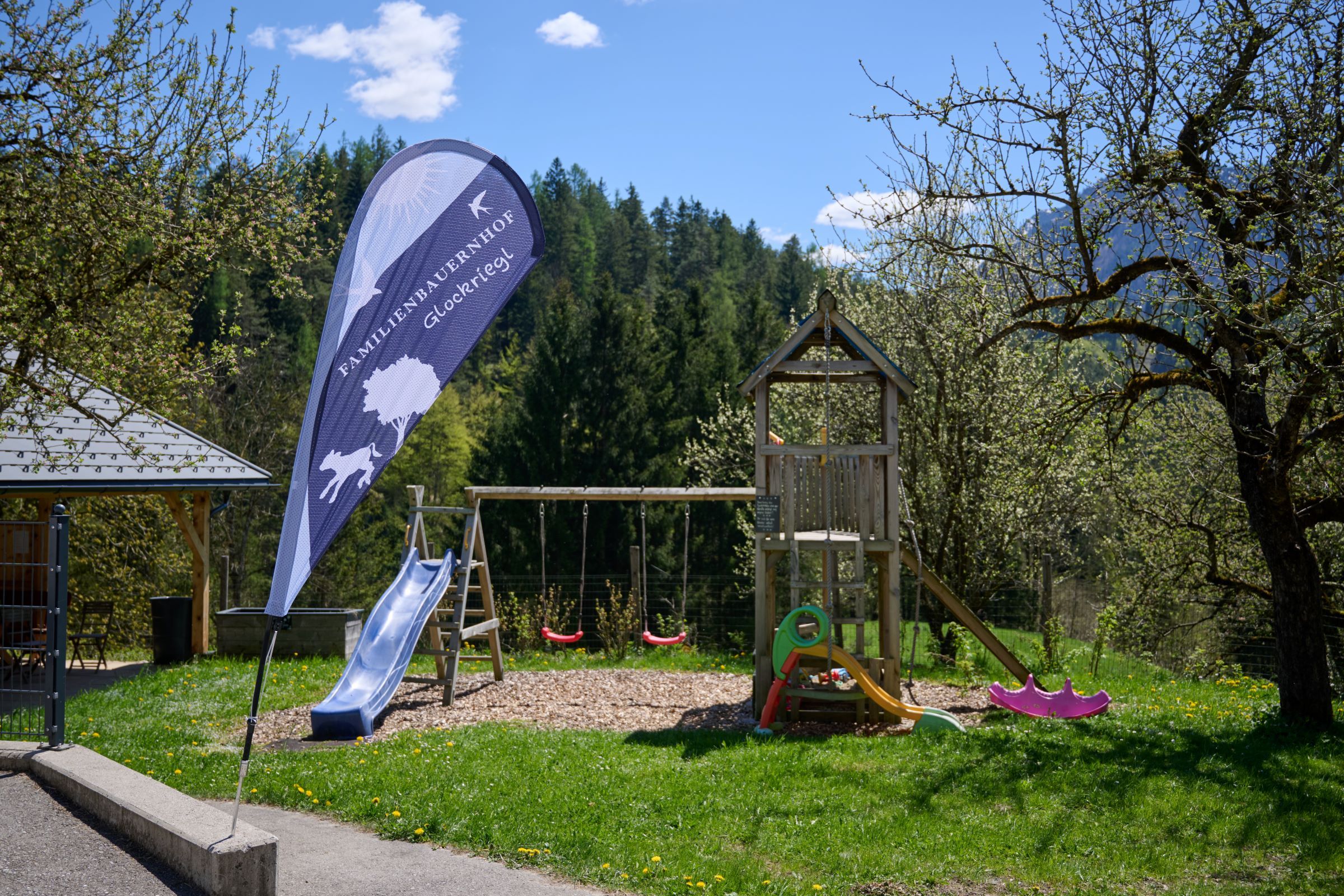 Spielplatz mit Rutsche und Schaukeln, umgeben von Bäumen und Natur.