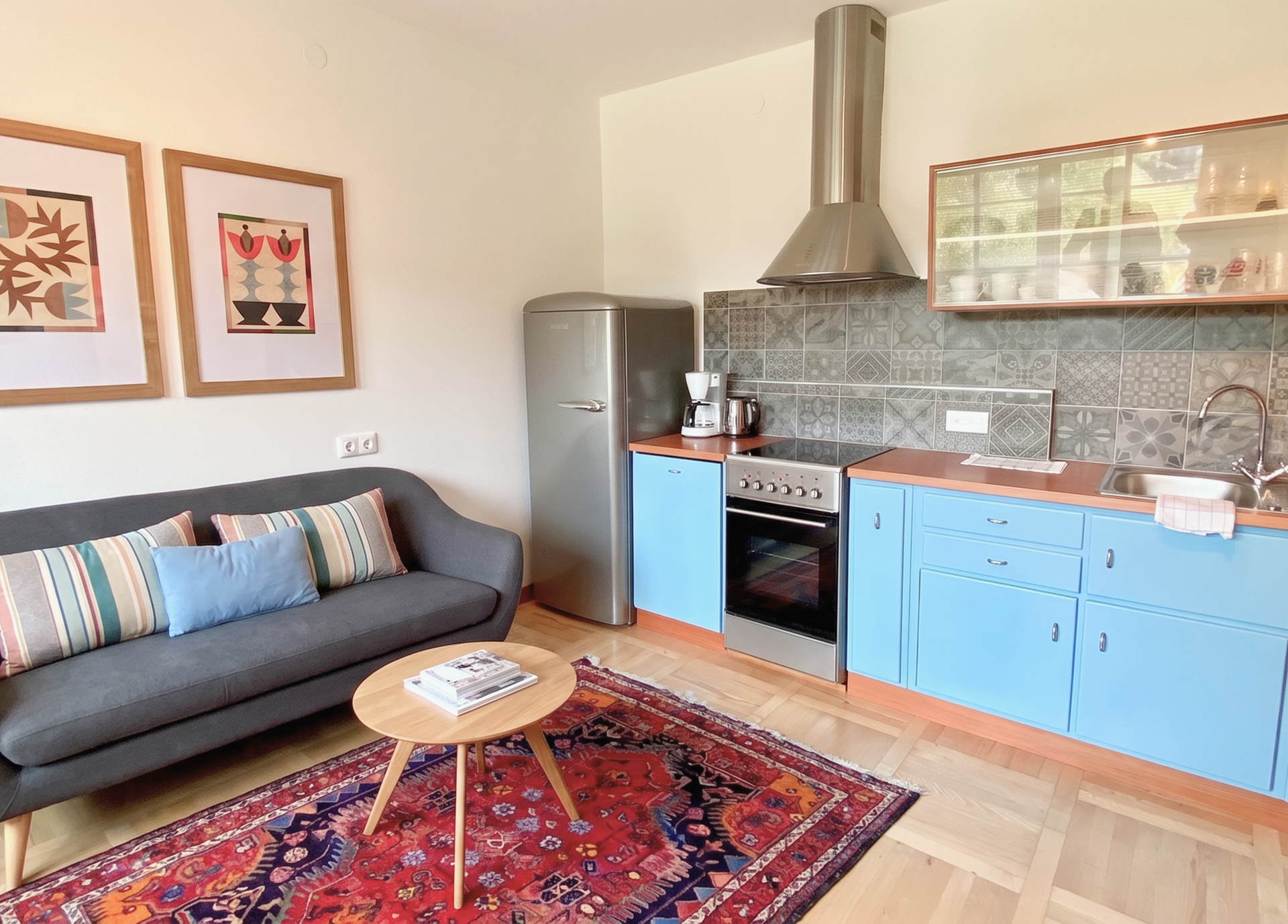 Gemütliche Küche mit blauen Schränken, grauem Sofa und buntem Teppich.