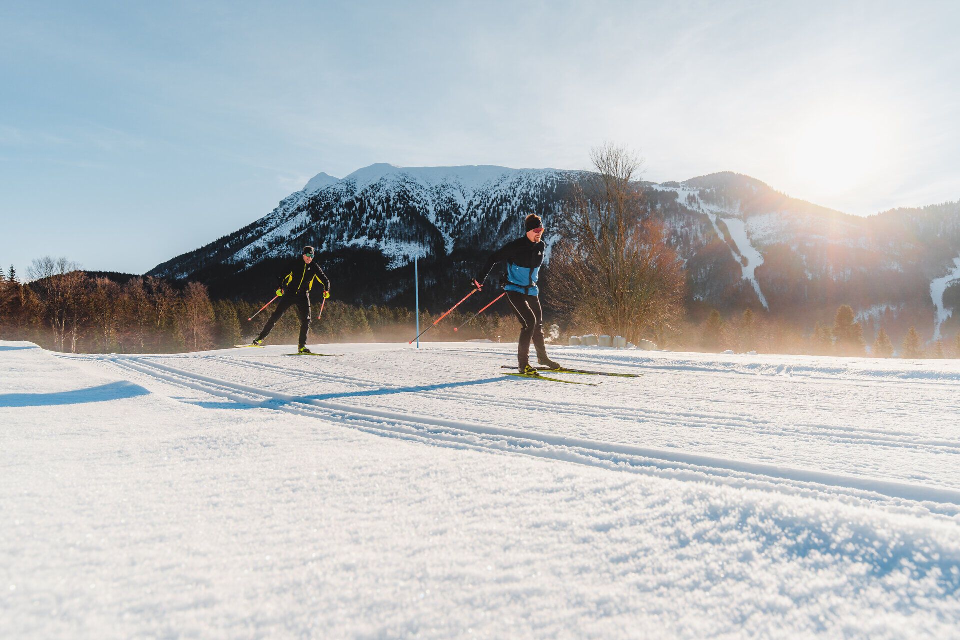 Die glitzernde Schneedecke erstreckt sich unter einem strahlend blauen Himmel, während zwei Langläufer elegant durch die winterliche Landschaft gleiten. Die majestätischen Berge im Hintergrund verleihen der Szenerie eine beeindruckende Kulisse und laden zu unvergesslichen Abenteuern in der Natur ein.