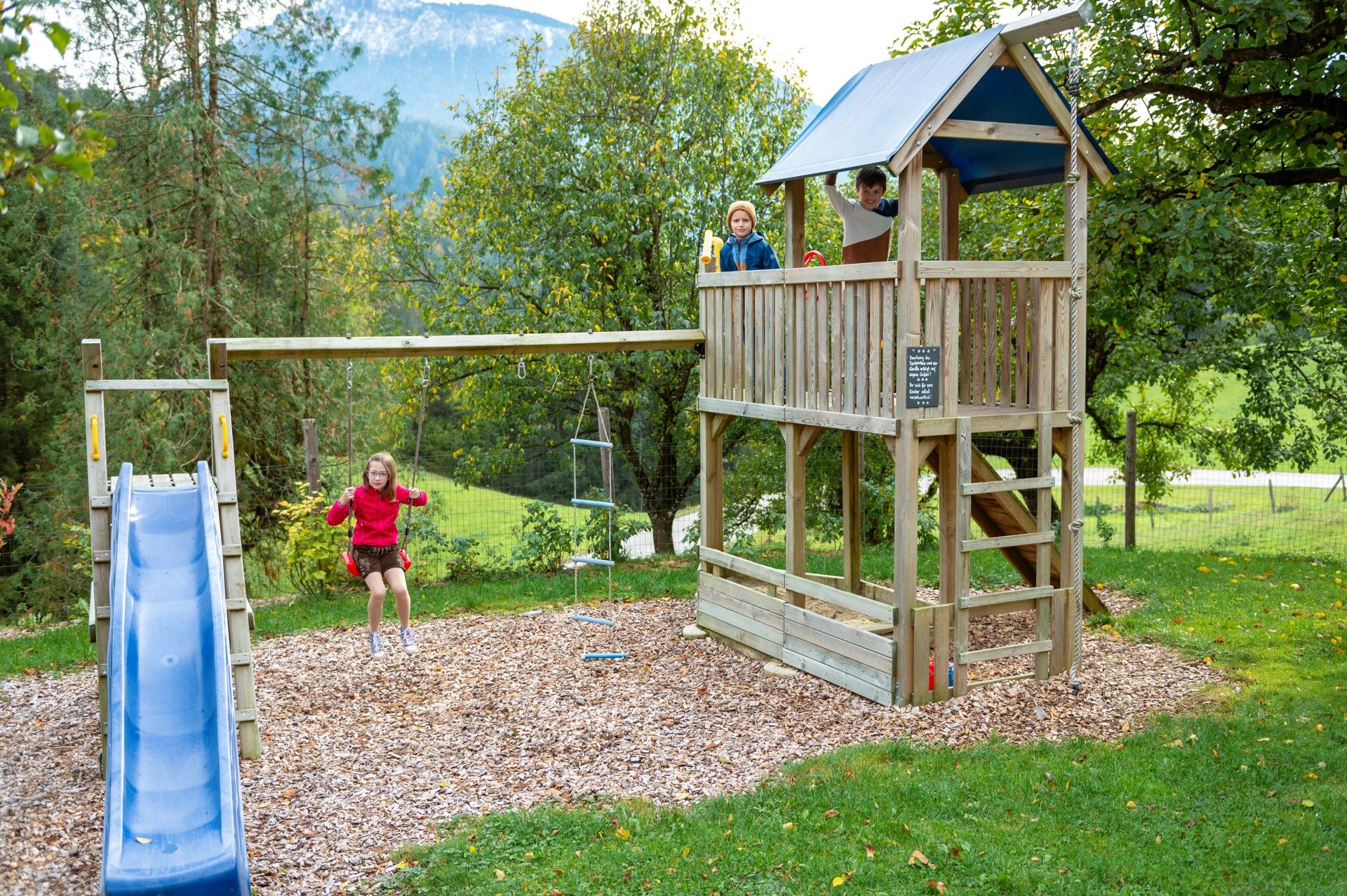 Kinder spielen auf einem Spielplatz mit Schaukel, Rutsche und Kletterturm in einer grünen Umgebung.