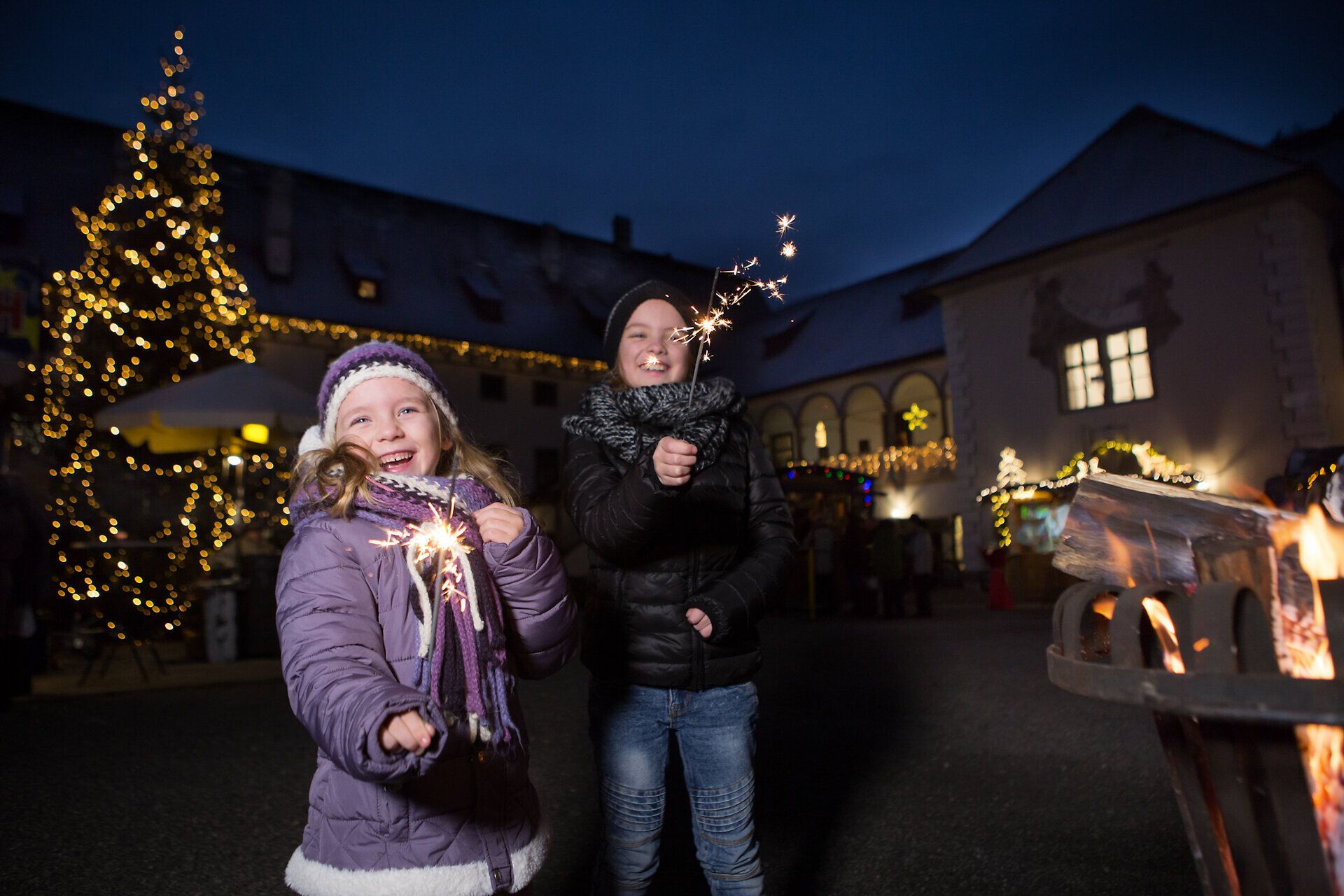 Zwei fröhliche Kinder strahlen mit funkelnden Wunderkerzen in der Hand, während die festliche Atmosphäre des Weihnachtsmarktes sie umgibt. Der glitzernde Weihnachtsbaum im Hintergrund und die sanften Lichter schaffen eine magische Stimmung, die die Vorfreude auf das Fest spürbar macht.