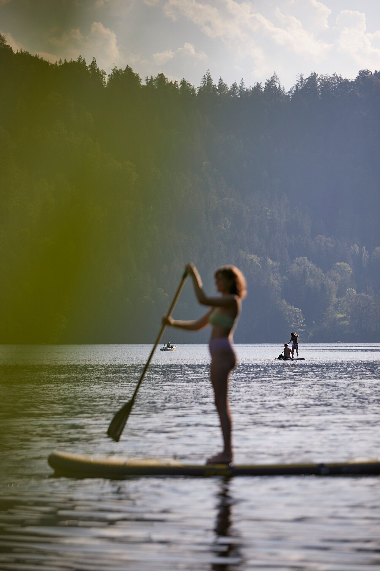 Sanfte Wellen glitzern im Sonnenlicht, während eine Person auf einem Stand-Up-Paddleboard über den ruhigen See gleitet. Umgeben von majestätischen Bergen und üppigem Grün, vermittelt die Szenerie ein Gefühl von Freiheit und Abenteuer.