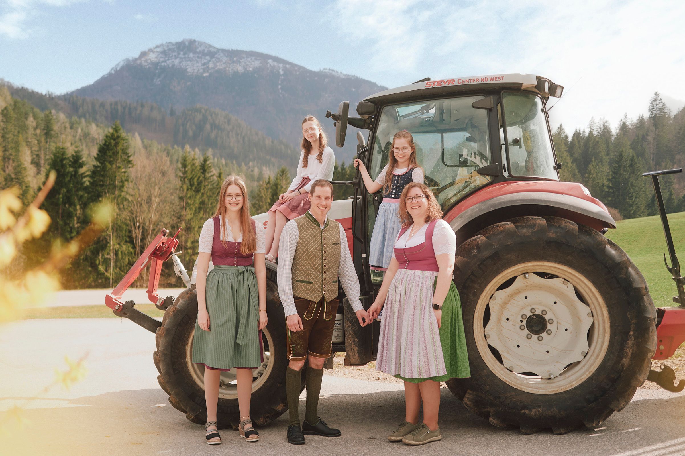 Eine Familie in traditioneller Tracht steht vor einem Traktor in einer ländlichen Berglandschaft.