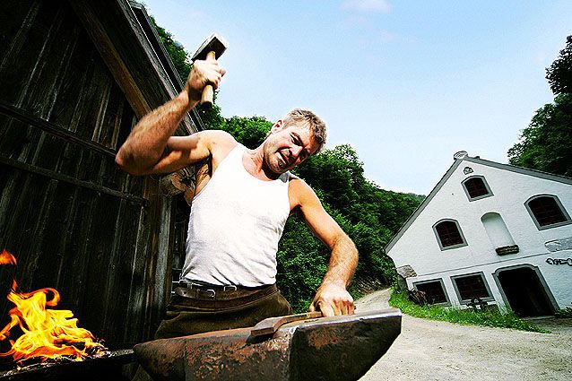 Ein Schmied arbeitet mit einem Hammer auf einem Amboss vor einem Gebäude im Freien.