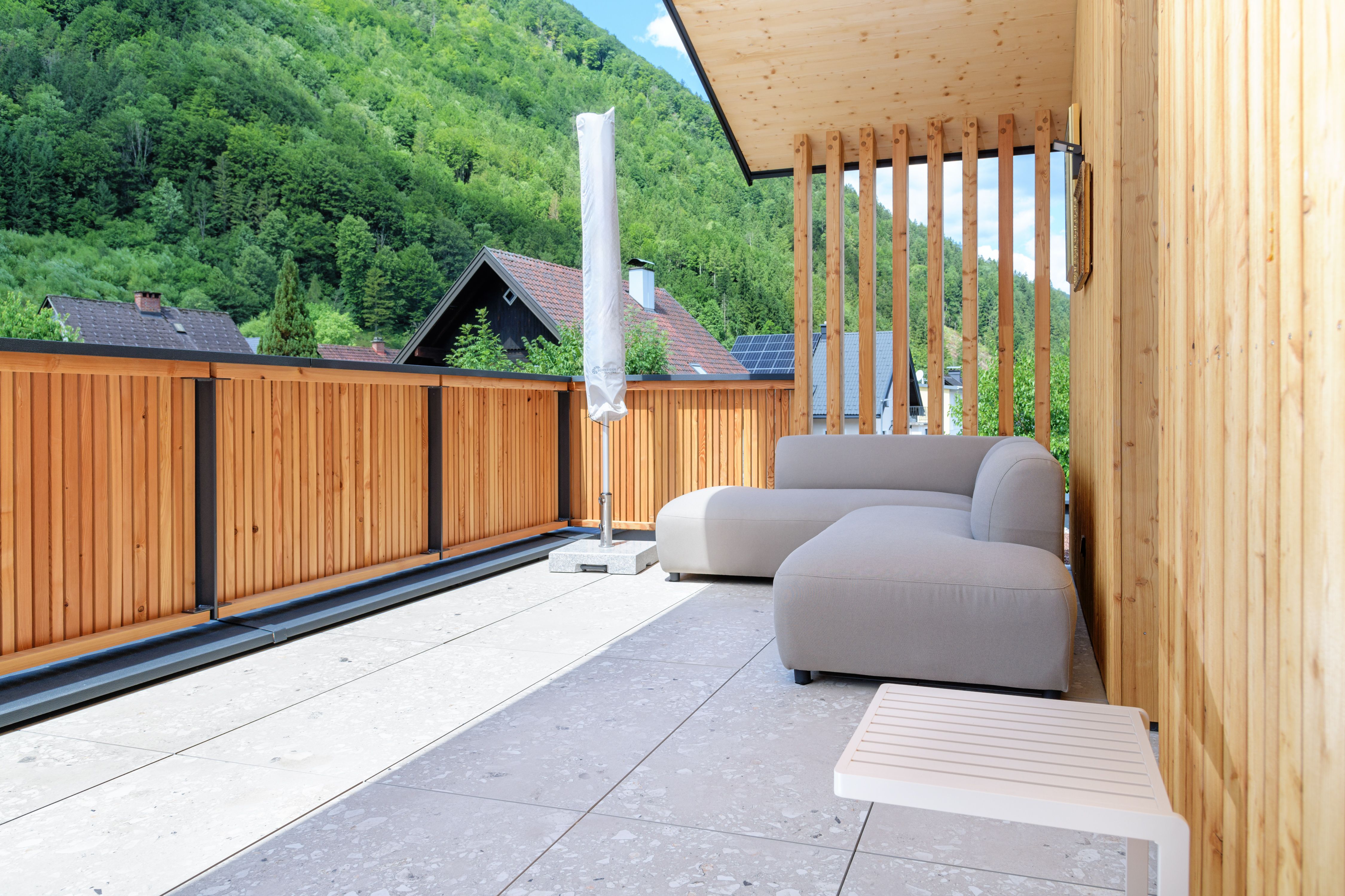 Moderne Terrasse mit Holzwänden, grauem Sofa und Blick auf bewaldete Hügel.