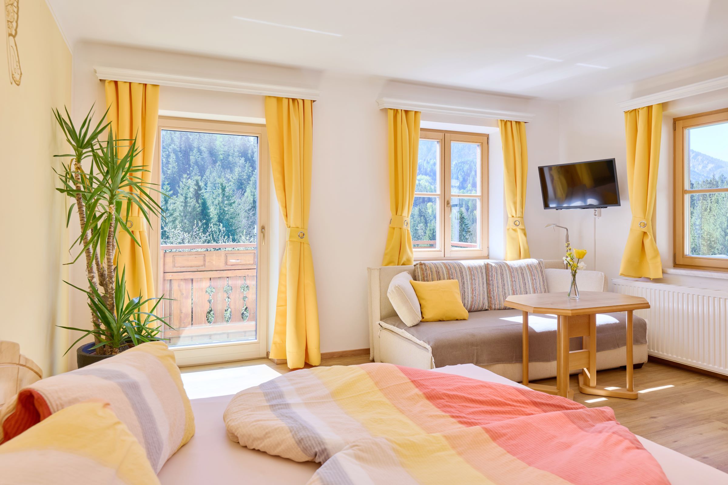 Heller Raum mit Bett, Sofa, Tisch und gelben Vorhängen, Blick auf Wald und Berge.