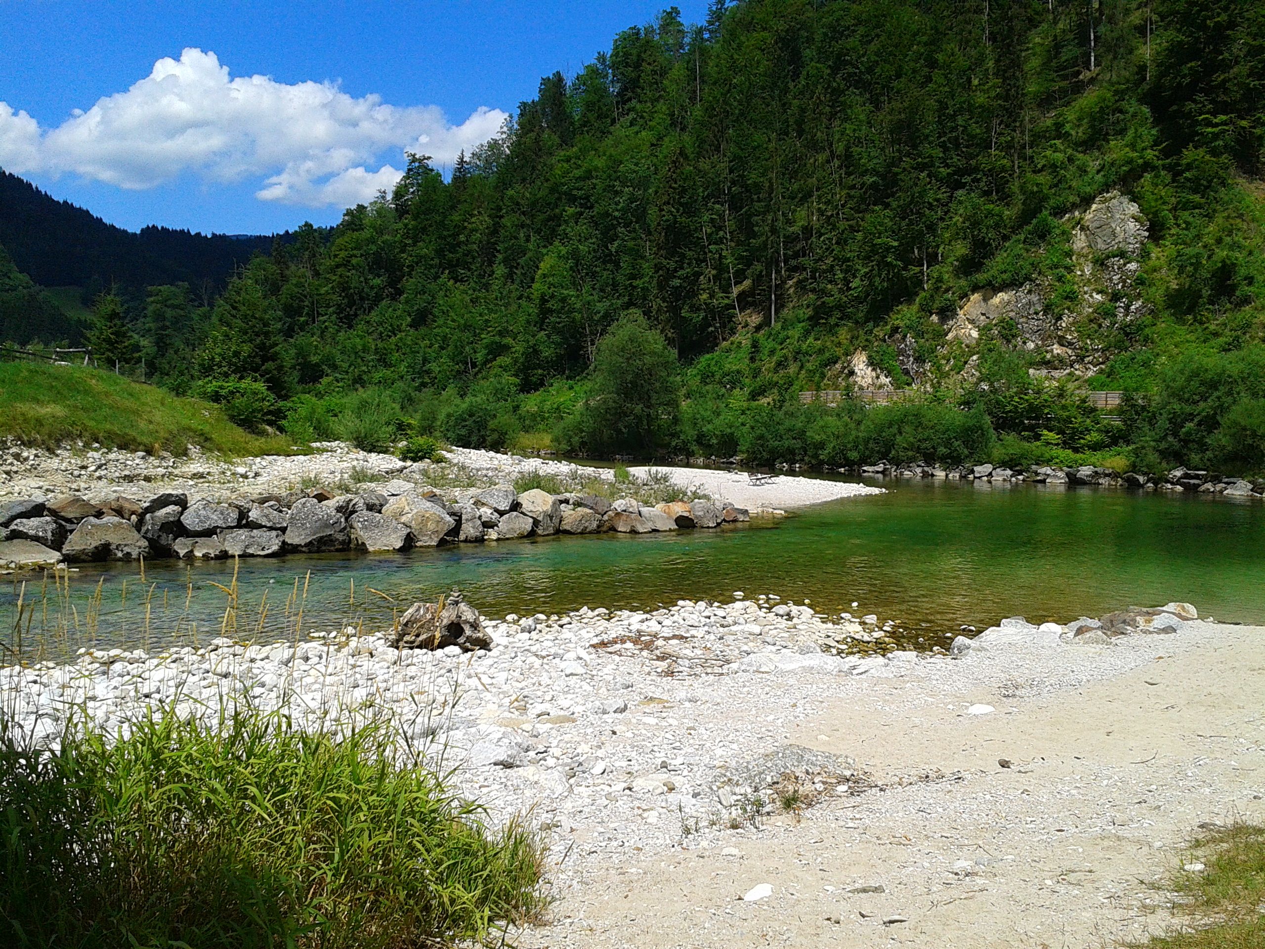 Flusslandschaft mit klarem Wasser, Kieselstrand und bewaldeten Hügeln.