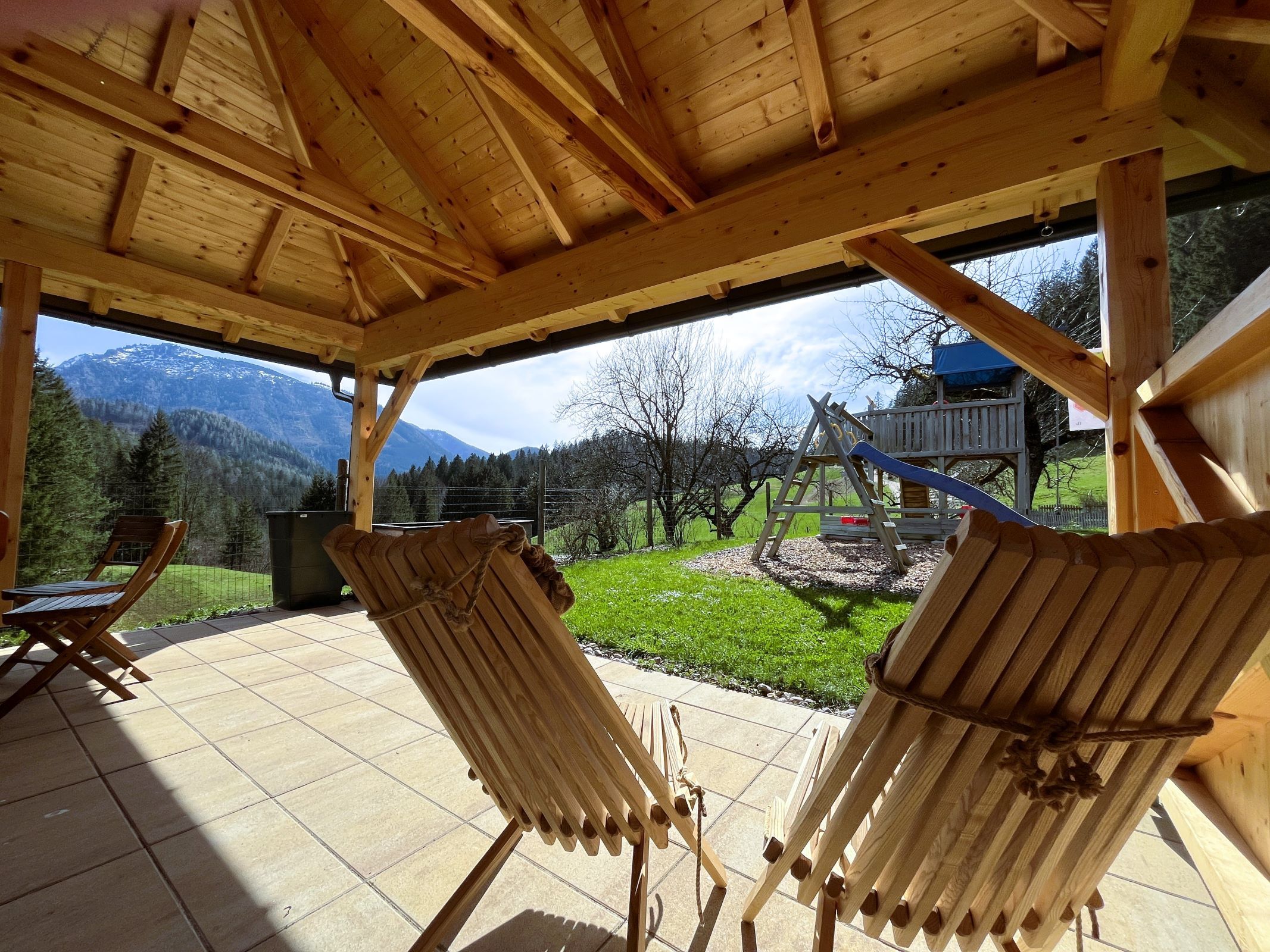 Blick aus einem Holzpavillon auf eine Berglandschaft mit Schaukel und Rutsche im Garten.