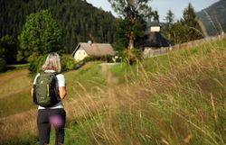 Pilgerin auf steilem Wiesenhang mit Blick auf Bauernhof.