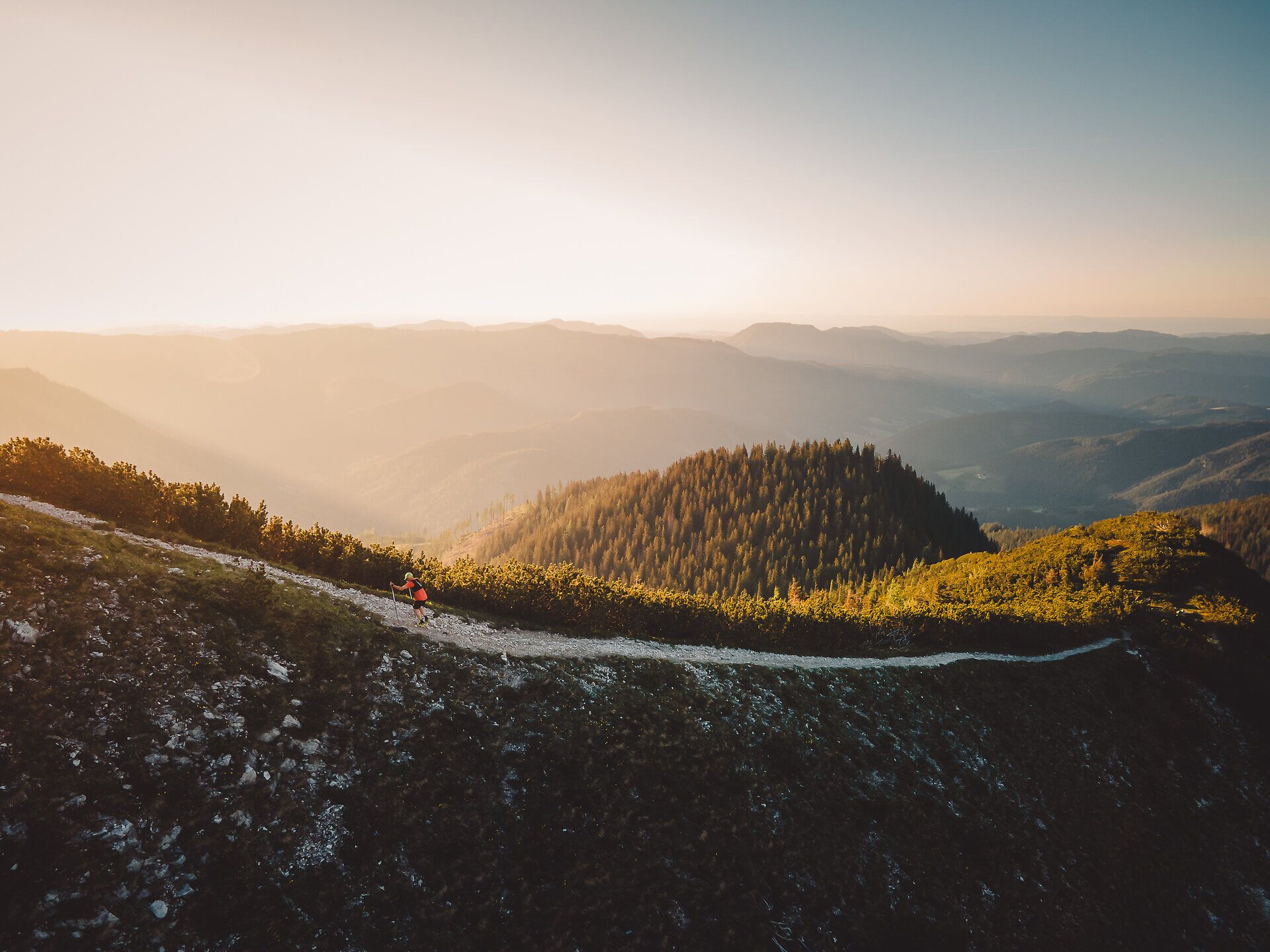Ein Trailläufer läuft auf einem schmalen Steinweg in der Abendsonne mit Blick in die umliegenden Bergwelt.