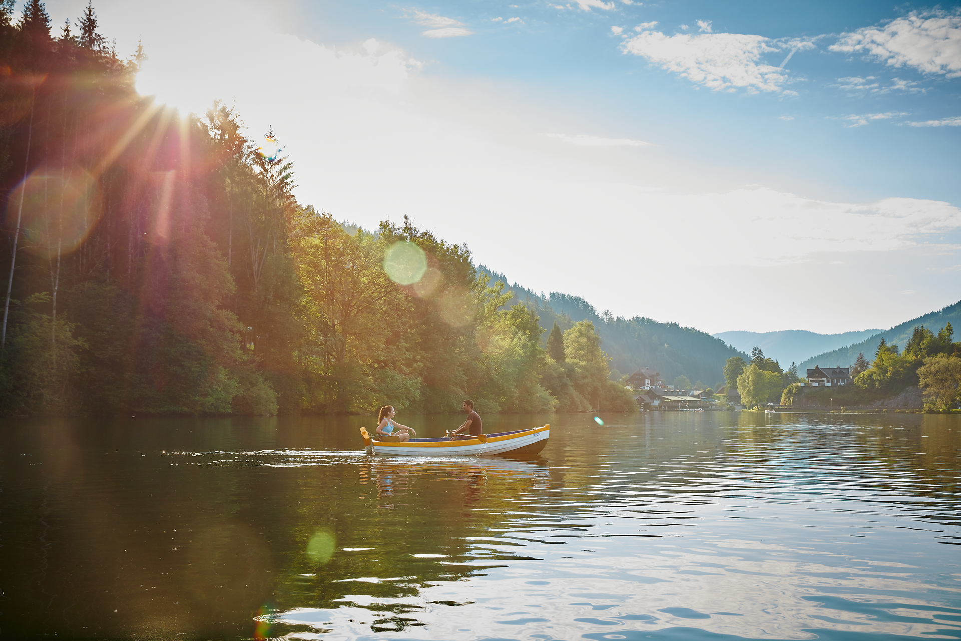 Die sanften Wellen des Sees spiegeln die strahlende Sonne wider, während zwei Personen in einem bunten Boot gemächlich über das Wasser gleiten. Umgeben von üppigem Grün und majestätischen Bergen, lädt diese idyllische Szenerie dazu ein, die Ruhe und Schönheit der Natur zu genießen.