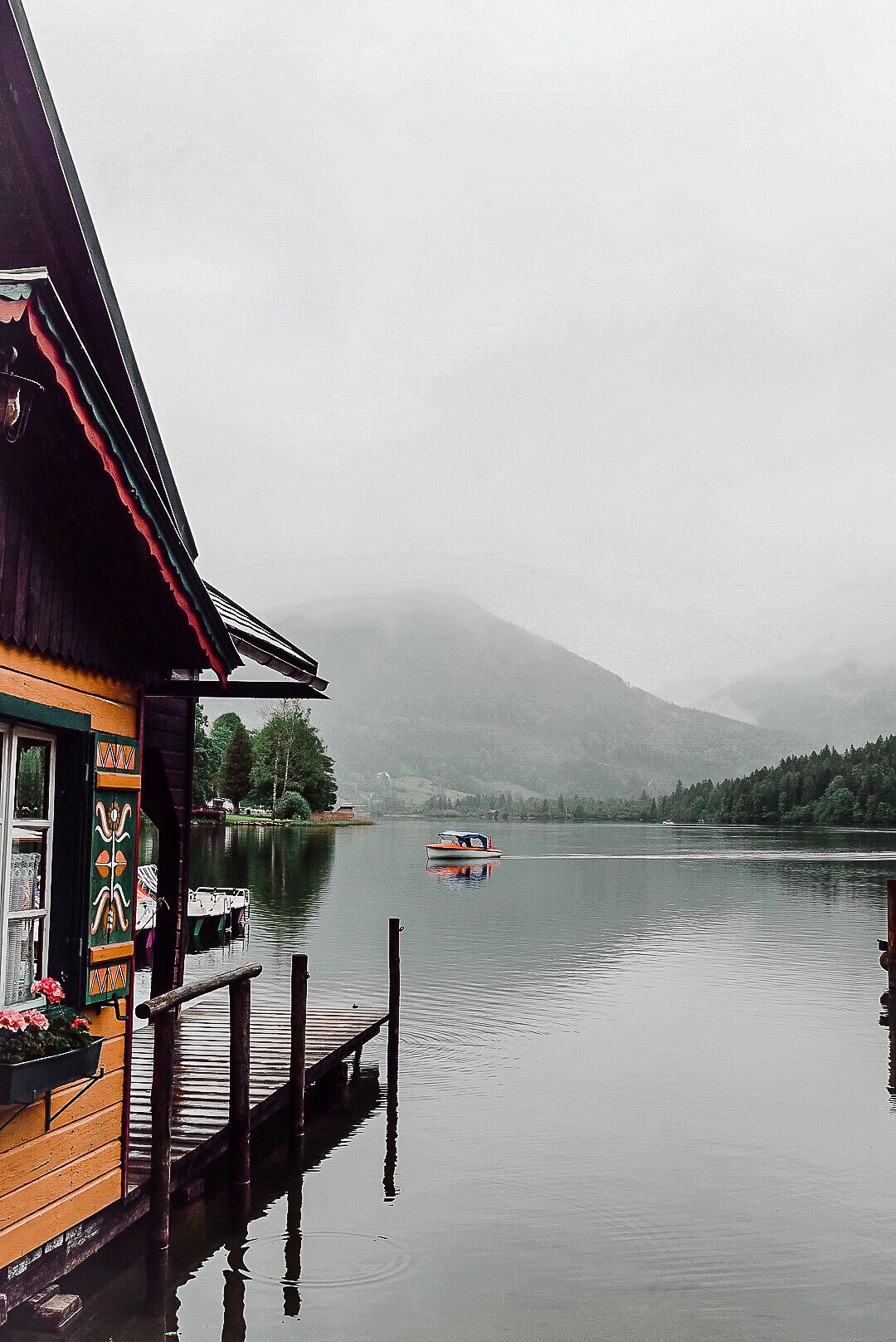 Ein sanfter Nebel umhüllt die ruhige Wasseroberfläche, während die bunten Fensterläden des charmanten Hauses am Ufer einen einladenden Kontrast zur grauen Umgebung bieten. Die majestätischen Berge im Hintergrund versprechen unvergessliche Abenteuer in der Natur.