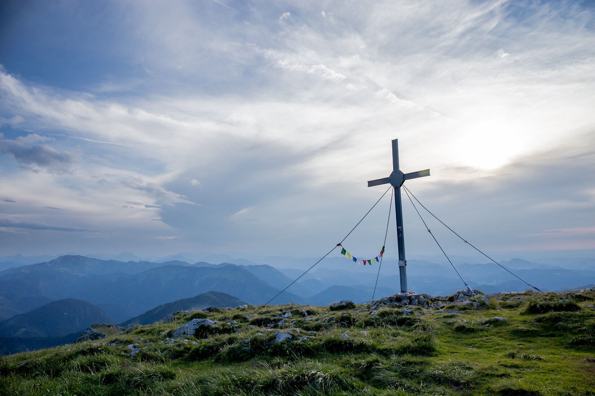 Ein majestätisches Gipfelkreuz thront über den sanften Hügeln, umgeben von einer atemberaubenden Berglandschaft. Die Wolken ziehen sanft über den Himmel und verleihen der Szenerie eine friedliche Atmosphäre, während die frische Bergluft zum Wandern einlädt.