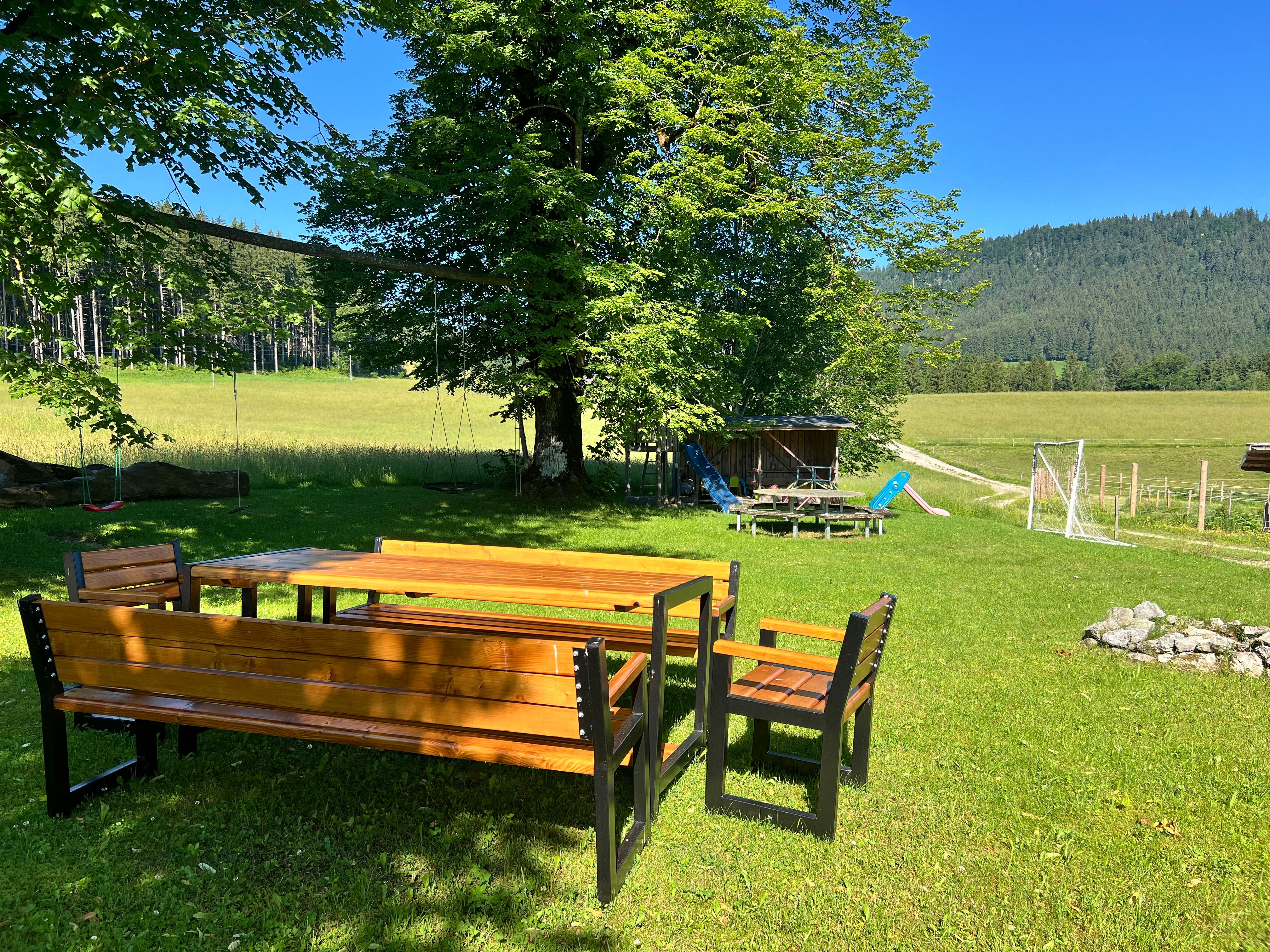 Ein Garten mit Holztisch und Bänken, umgeben von Bäumen und Wiesen, mit einem Spielplatz im Hintergrund.