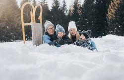 Eine fröhliche Familie genießt den Winter in der schneebedeckten Landschaft. Die Kinder lachen und spielen im Schnee, während die Erwachsenen entspannt im Hintergrund liegen. Die strahlende Sonne und die majestätischen Ybbstaler Alpen schaffen eine zauberhafte Atmosphäre.
