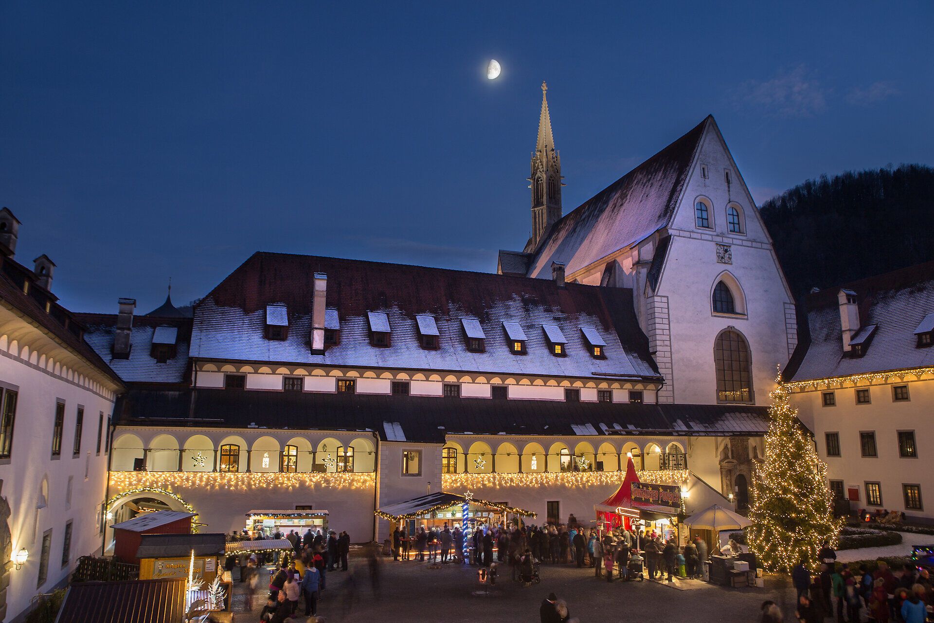 In der zauberhaften Kulisse des Adventmarktes erstrahlen festlich geschmückte Stände und ein prächtiger Weihnachtsbaum. Die sanfte Beleuchtung und der klare Nachthimmel mit einem leuchtenden Mond schaffen eine stimmungsvolle Atmosphäre, die die Herzen der Besucher erwärmt.