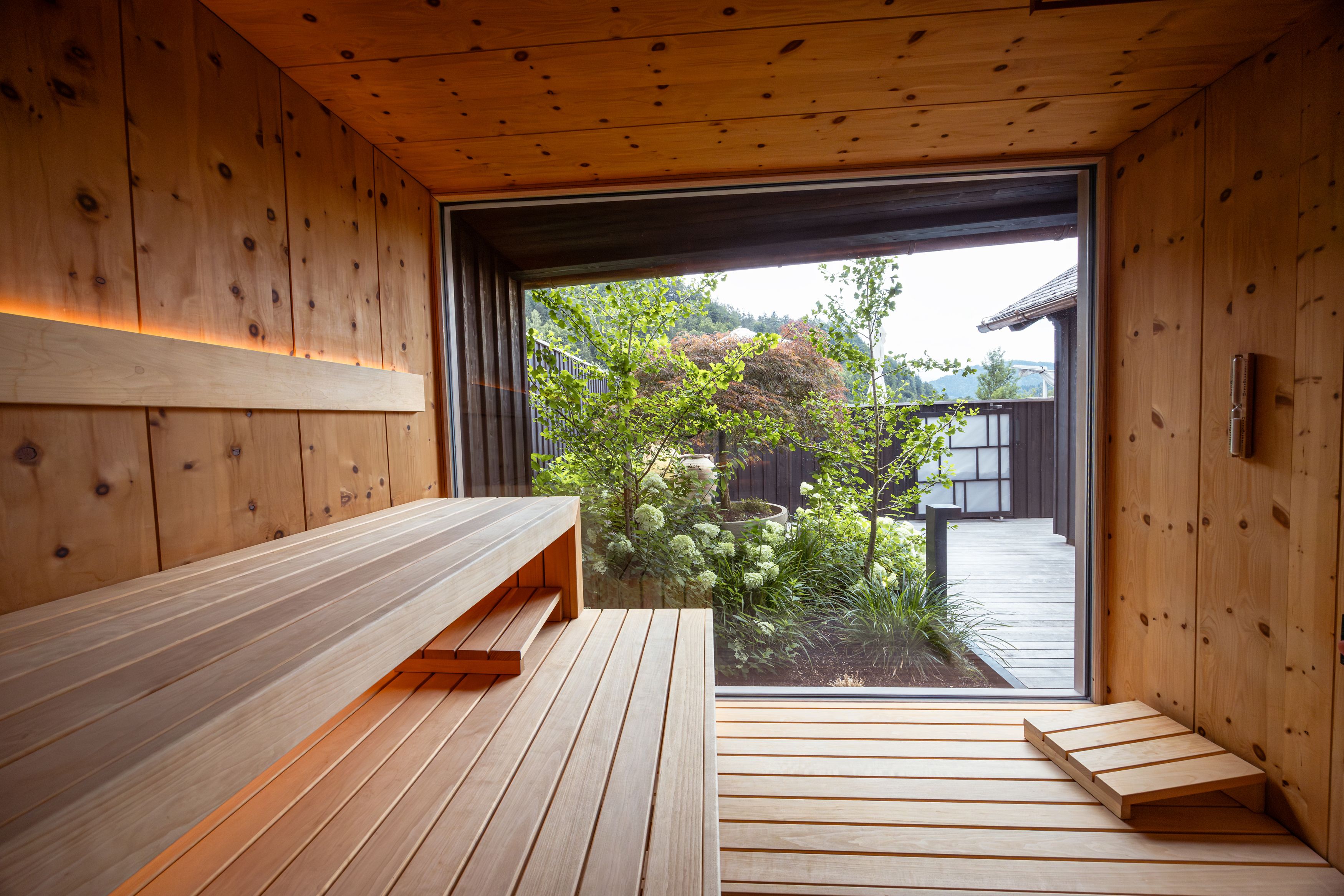 Innenansicht einer modernen Sauna mit Holzwänden und -bänken, Blick auf einen Garten durch ein großes Fenster.