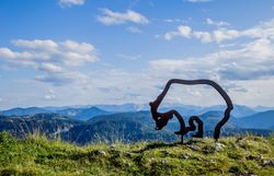 Ein majestätischer Blick auf die sanften Hügel und die beeindruckenden Berge, die sich unter einem strahlend blauen Himmel erstrecken. Die harmonische Verbindung von Natur und Kunst wird durch die stilvolle Skulptur des Ötscherbären unterstrichen, die den Wanderern ein Gefühl von Abenteuer und Freiheit vermittelt.