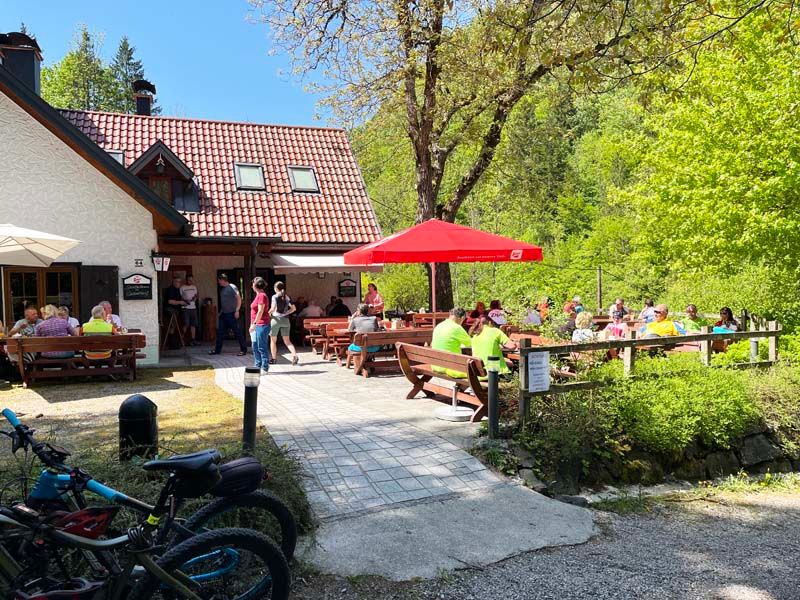 Eine Berghütte mit Terrasse, auf der Menschen an Tischen sitzen. Fahrräder sind im Vordergrund zu sehen, umgeben von grüner Natur.