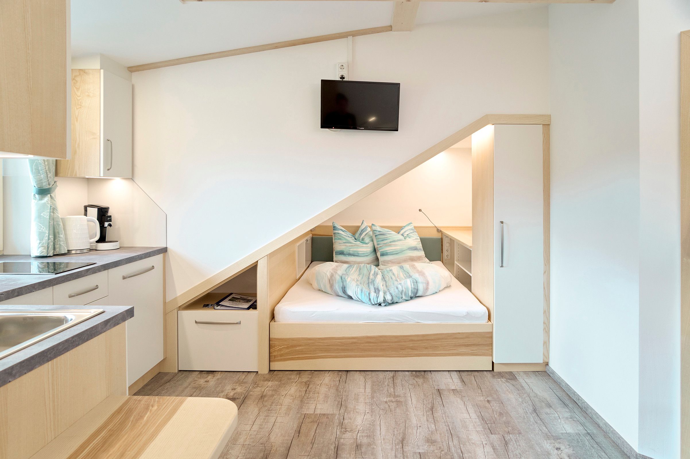 Kompaktes Apartment mit integriertem Bett unter einer Dachschräge, Küchenzeile und Fernseher.