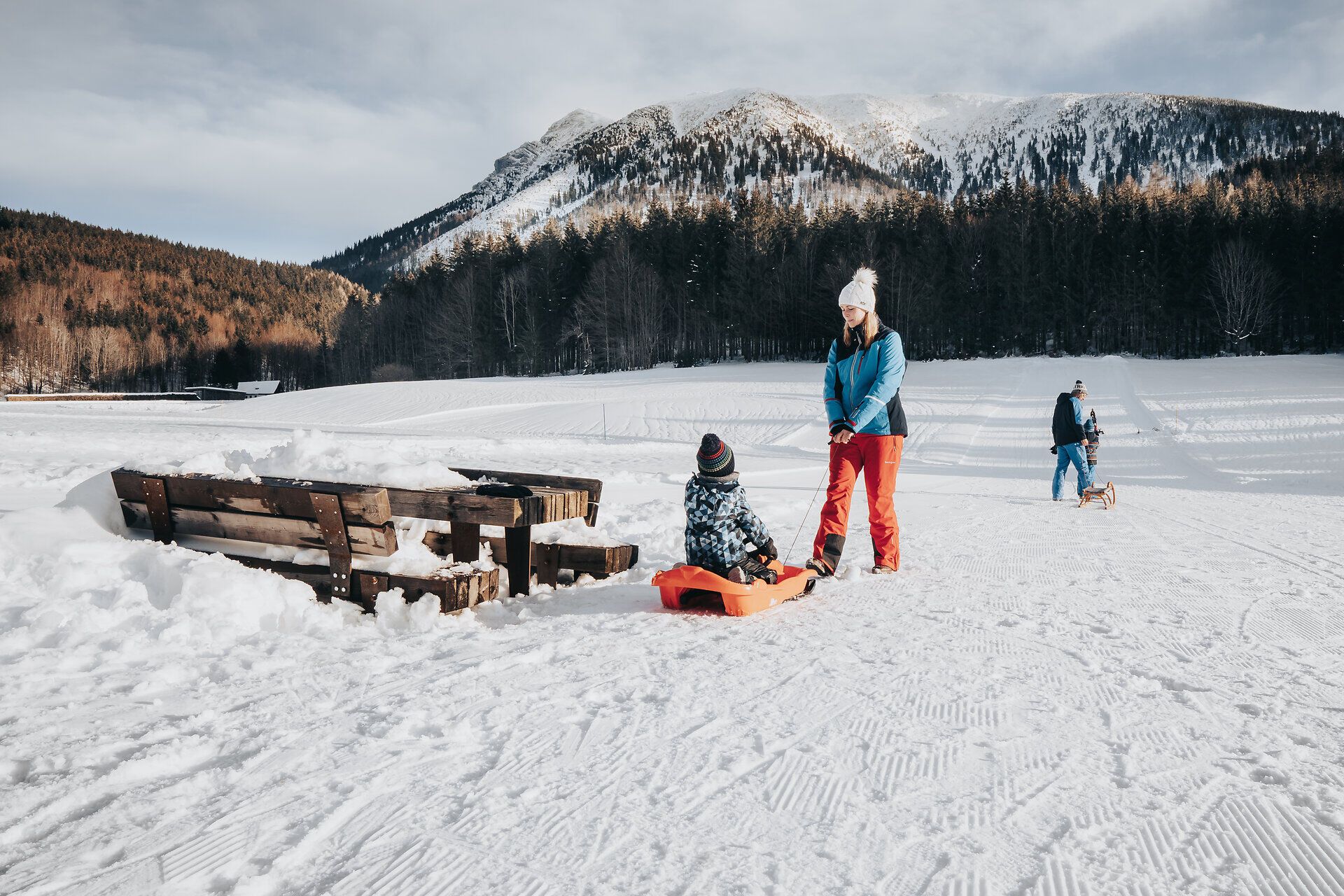 Eine Familie genießt den Winterzauber in den Ybbstaler Alpen, während ein Kind fröhlich auf einem Schlitten sitzt. Die schneebedeckte Landschaft strahlt Ruhe und Freude aus, während die Berge majestätisch im Hintergrund thronen. Hier wird der Spaß im Schnee zum unvergesslichen Erlebnis.