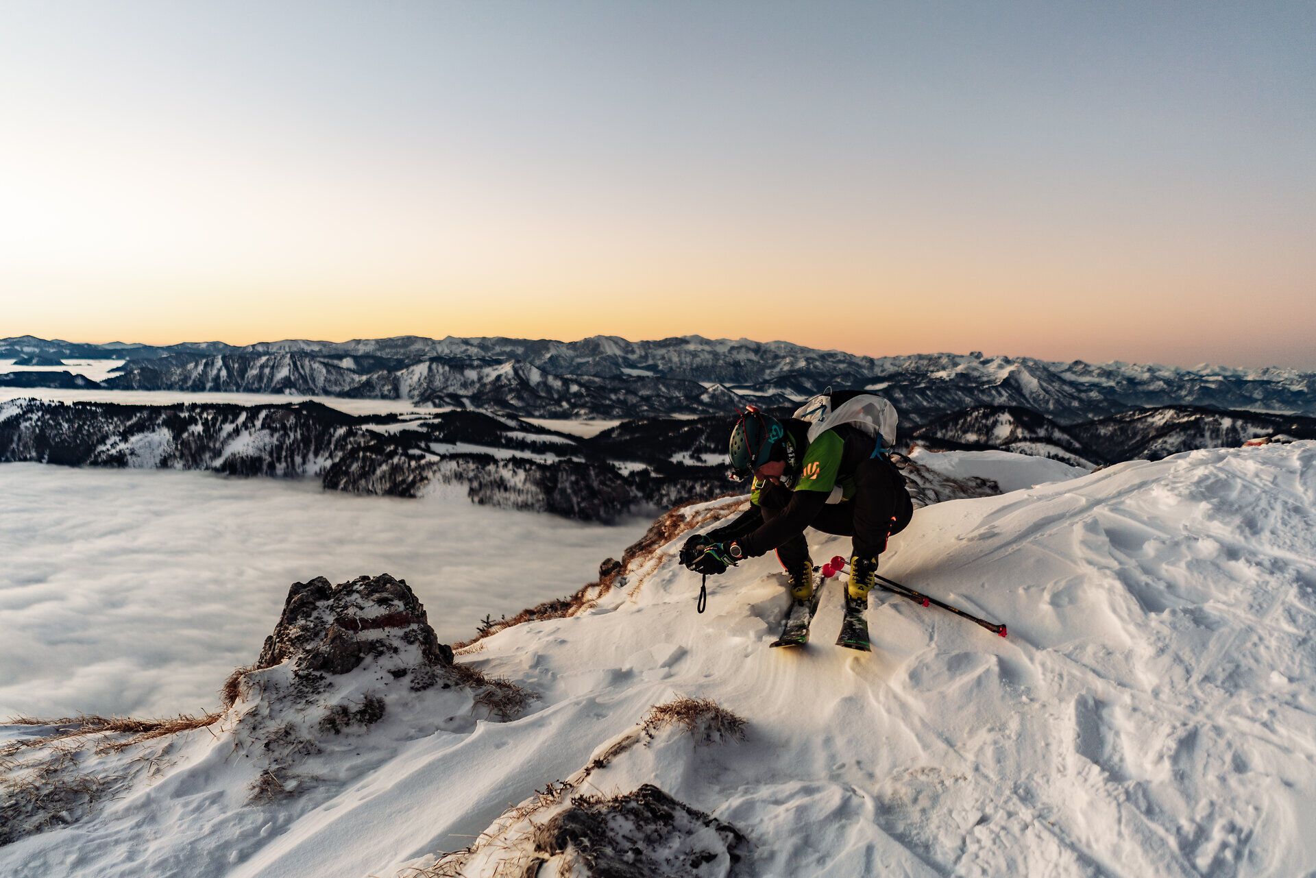 Skitourengeher kniet bei Sonnenaufgang auf einem verschneiten Berggrat und justiert seine Ausrüstung; im Hintergrund weite Winter-Berglandschaft über einem Meer aus Wolken.