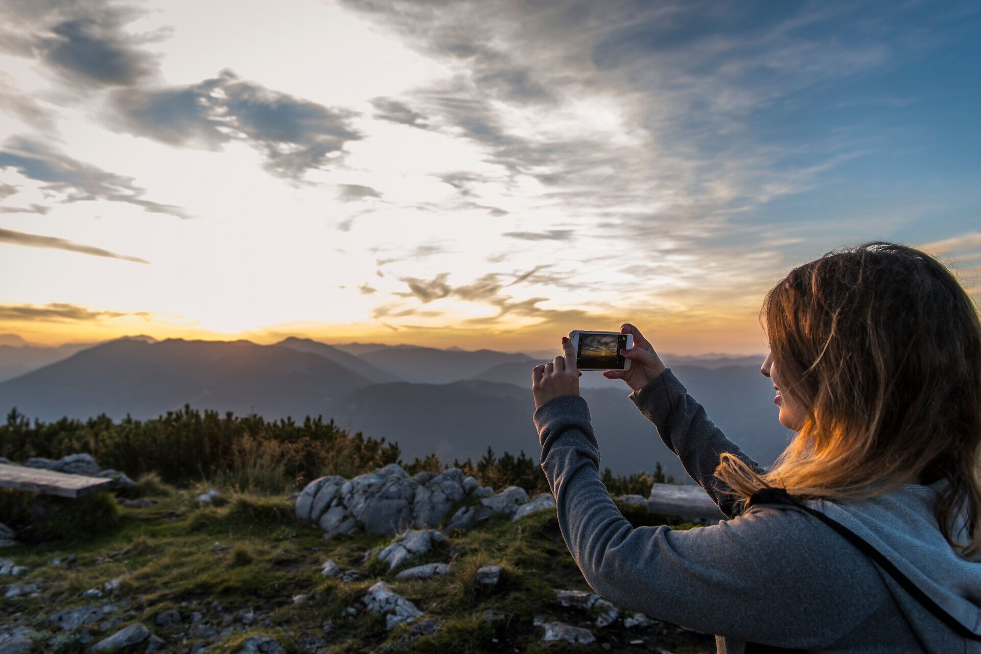 Ein atemberaubender Sonnenuntergang taucht die Berglandschaft in warmes Licht, während ein Wanderer den Moment mit seinem Handy festhält. Die sanften Hügel und majestätischen Gipfel laden dazu ein, die Schönheit der Natur zu genießen und die frische Bergluft zu atmen.