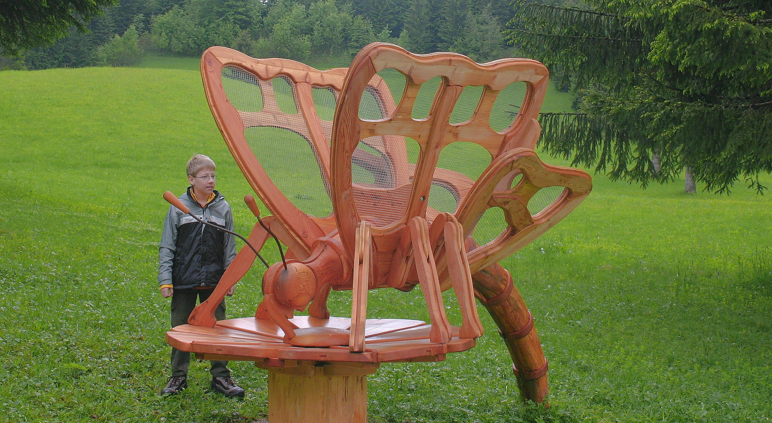 Ein Junge steht neben einer großen Holzskulptur eines Schmetterlings auf einer Wiese.