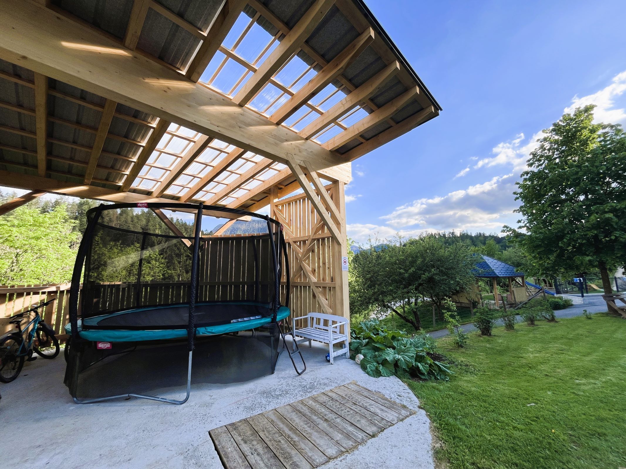 Überdachtes Trampolin in einem Garten mit Holzdach und umgebender Natur.