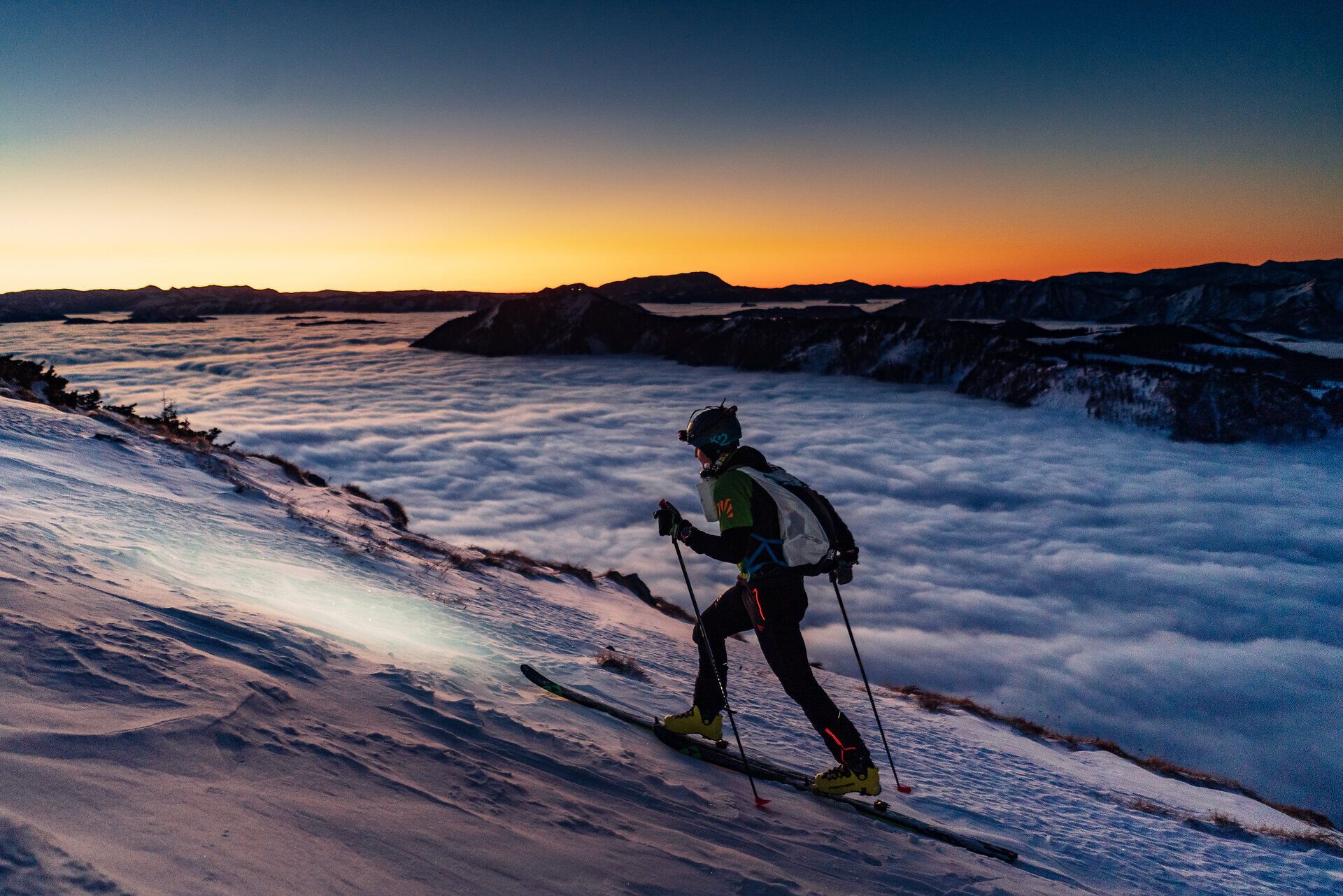 Skitourengeher vor Sonnenaufgang auf einem verschneiten Berggrat; im Hintergrund weite Winter-Berglandschaft über einem Meer aus Wolken.