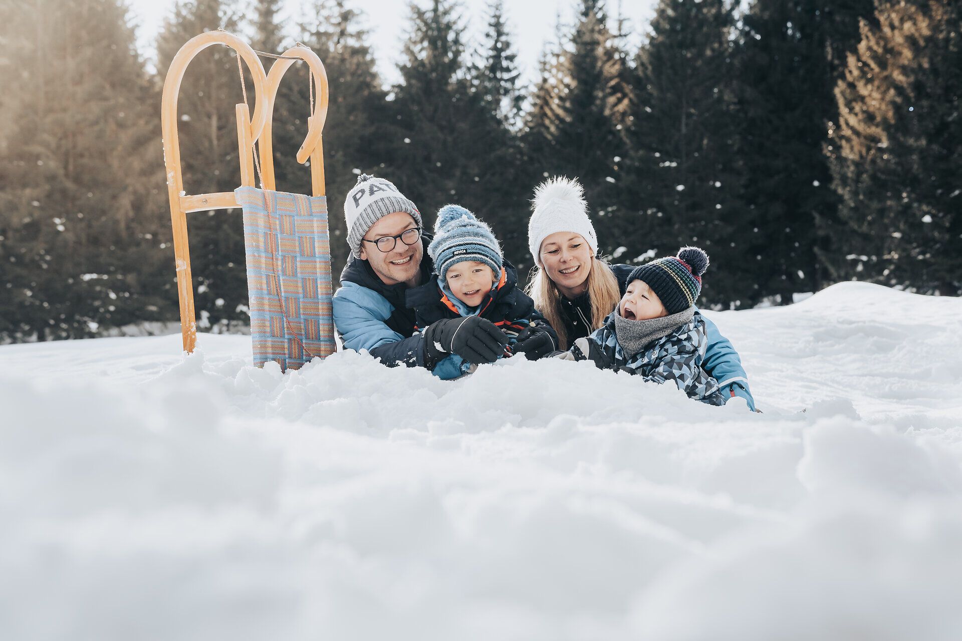 Eine fröhliche Familie genießt den Winter in der schneebedeckten Landschaft. Die Kinder lachen und spielen im Schnee, während die Erwachsenen entspannt im Hintergrund liegen. Die strahlende Sonne und die majestätischen Ybbstaler Alpen schaffen eine zauberhafte Atmosphäre.