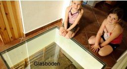 Zwei Kinder sitzen neben einem Glasboden, der einen Blick auf darunterliegende Strukturen ermöglicht.