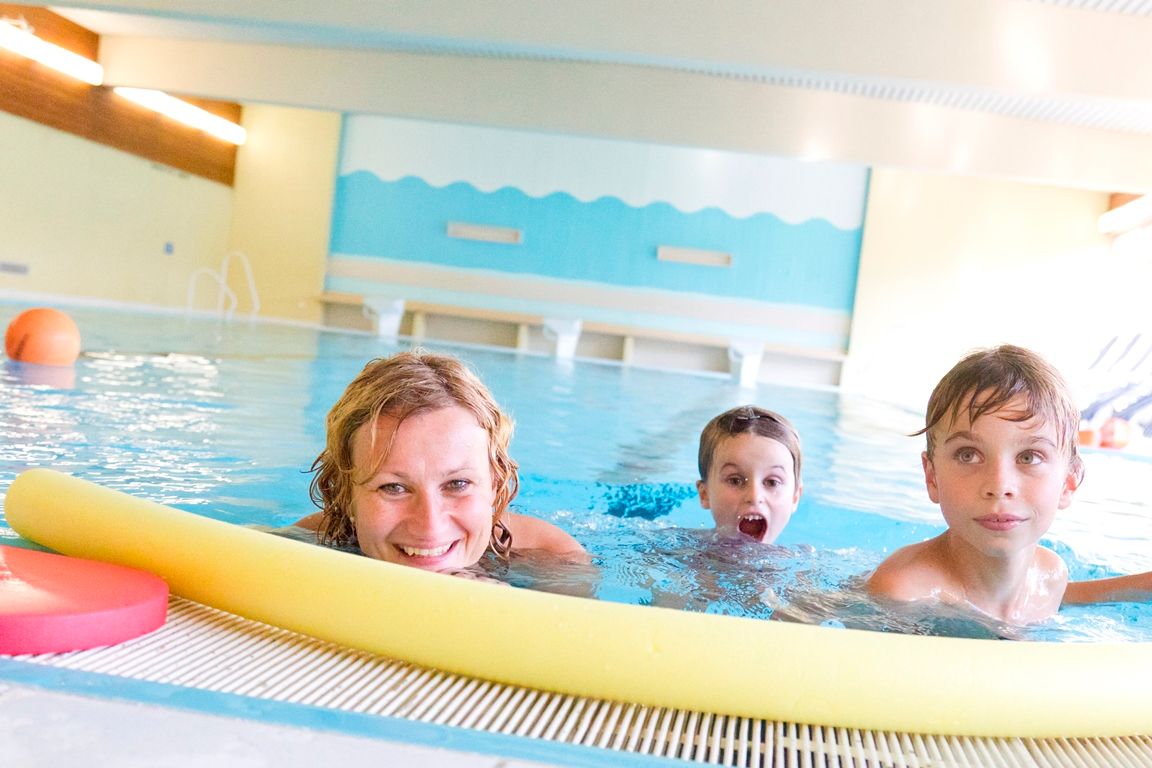 Eine Frau und zwei Kinder schwimmen in einem Hallenbad, lächelnd und spielend mit einer gelben Poolnudel.
