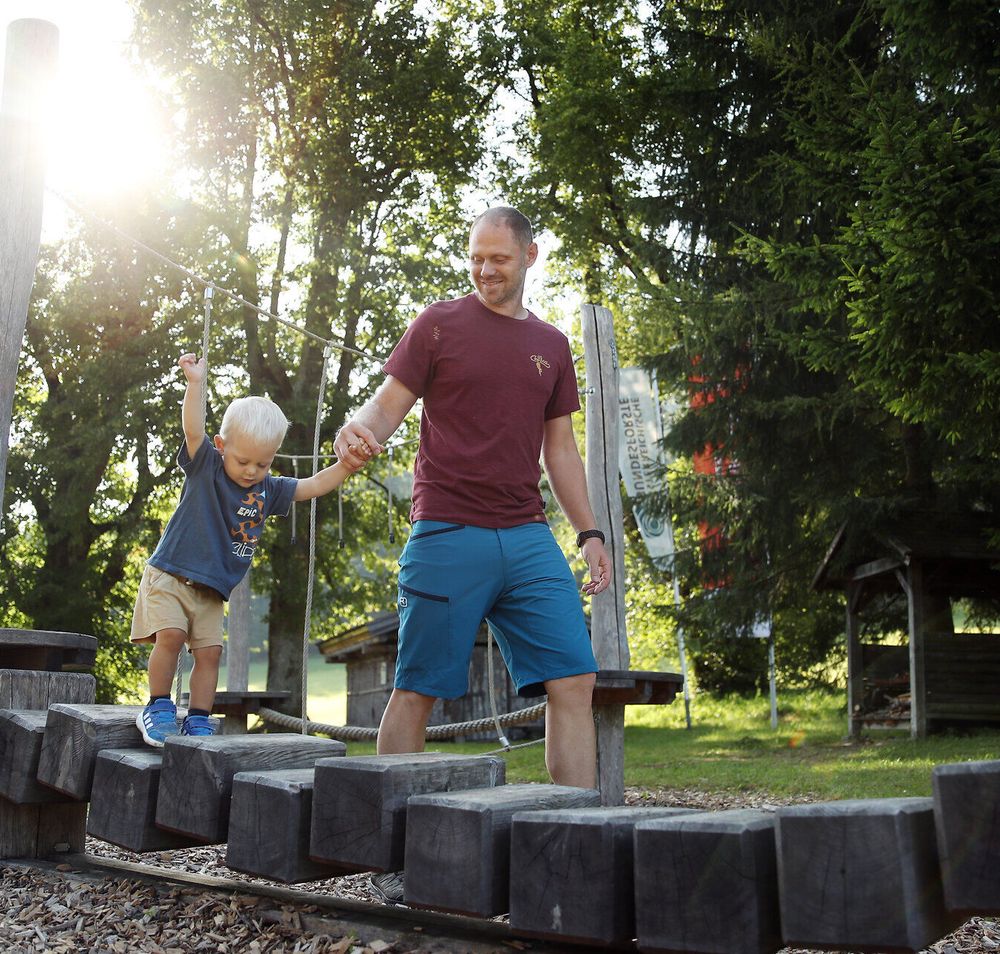 Ein Mann hält ein Kleinkind an der Hand während dieses über Holzblöcke am Spielplatz Promau balanciert. 