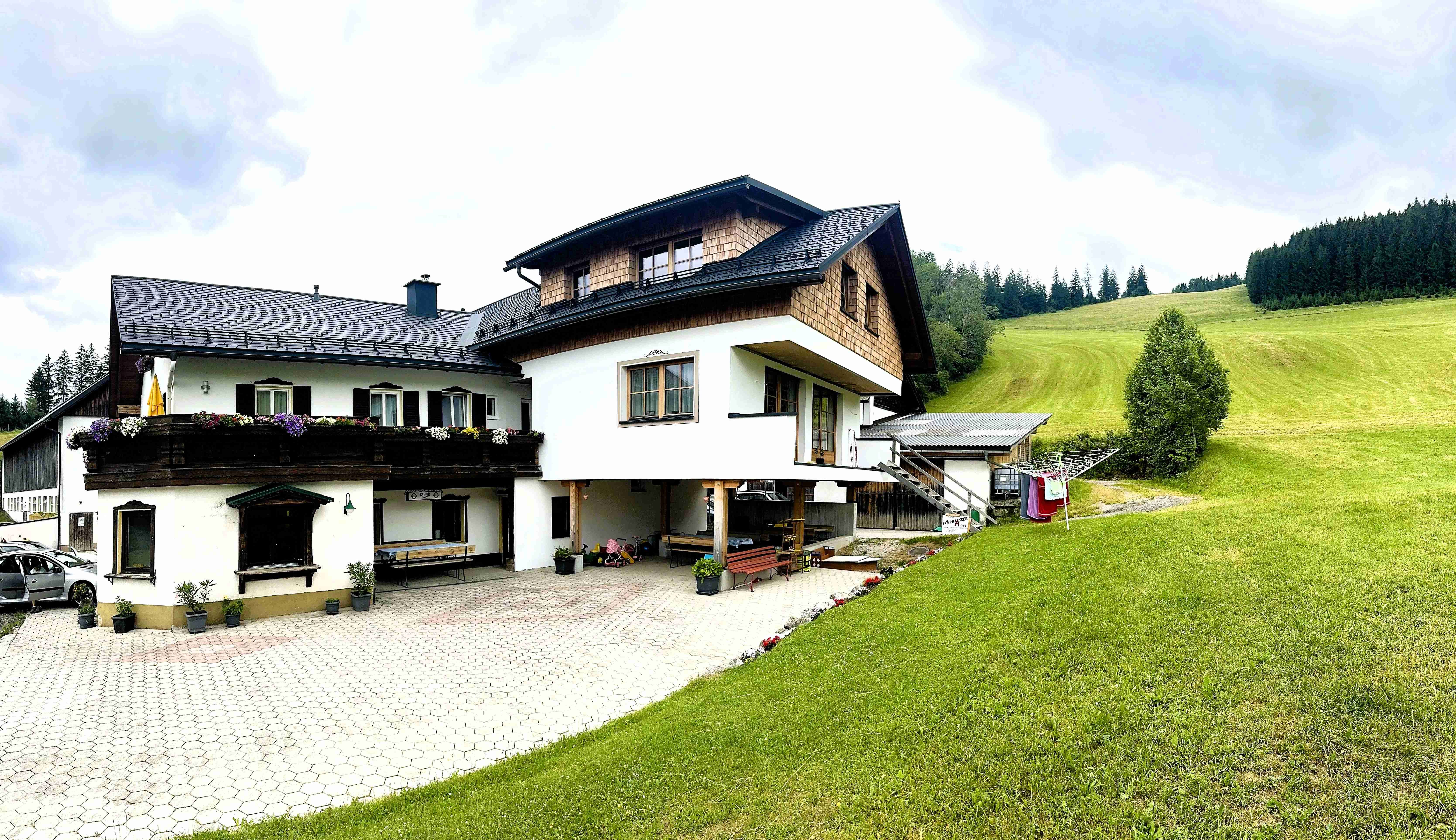 Ein traditionelles Bauernhaus mit Holzverkleidung und Blumen auf dem Balkon, umgeben von grünen Wiesen und Wäldern.