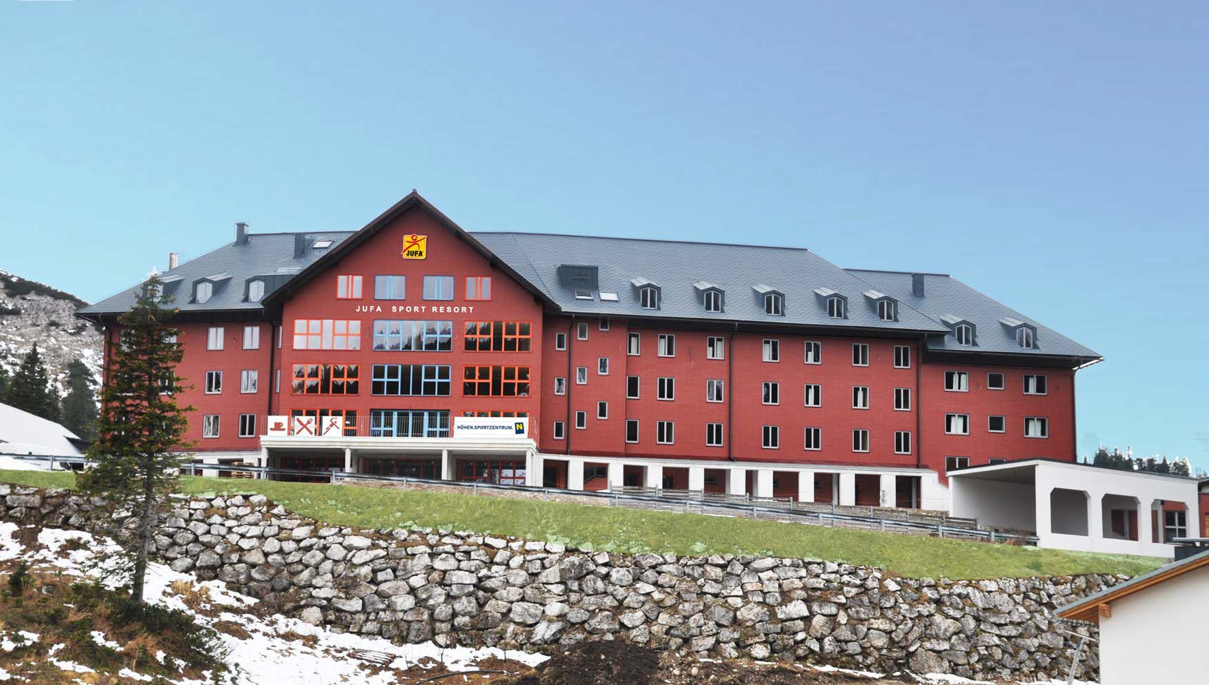 Großes rotes Gebäude des Jufa Sport Resorts in einer bergigen Landschaft.