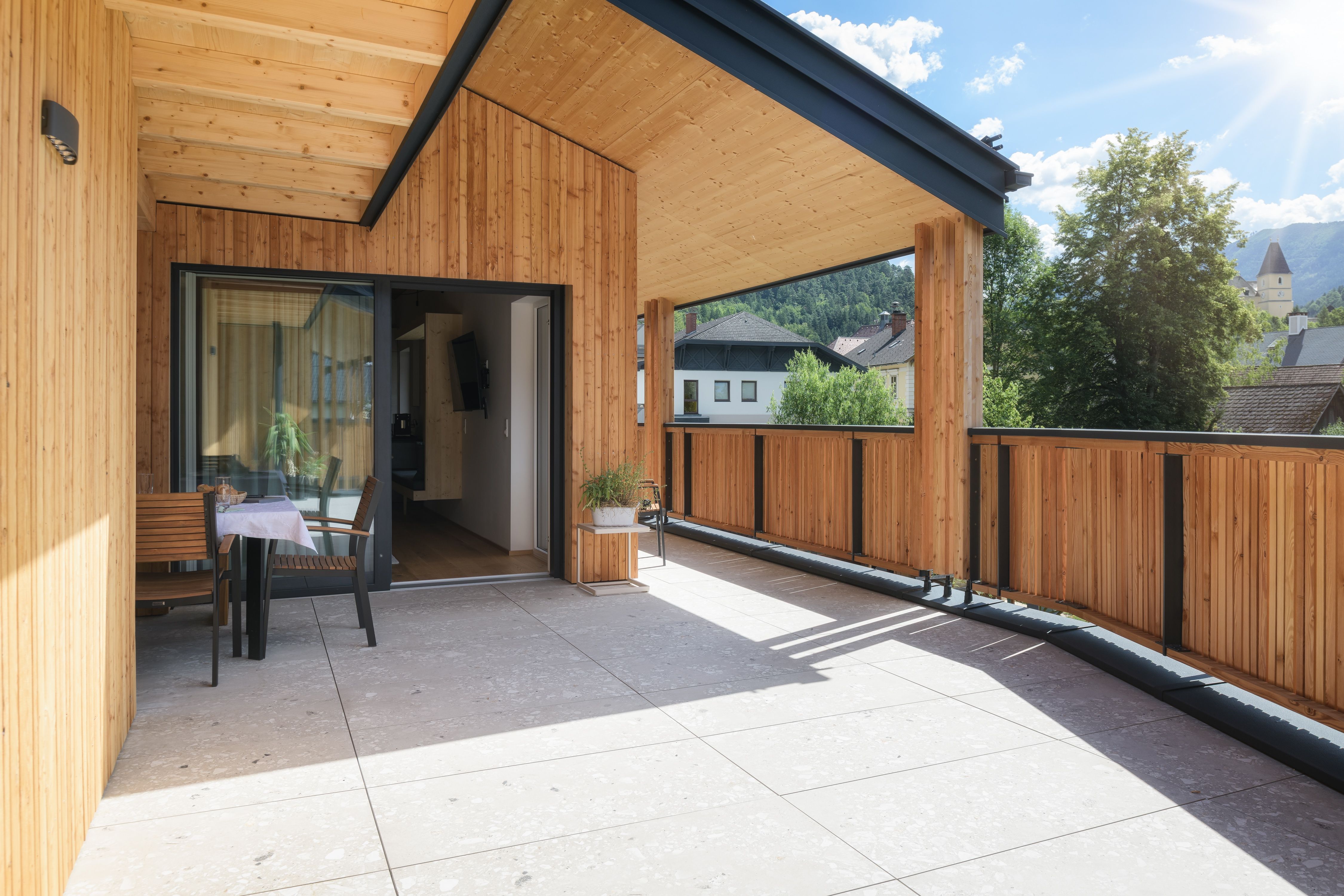 Moderne Holzterrasse mit Tisch und Stühlen, umgeben von Bäumen und Gebäuden im Hintergrund.