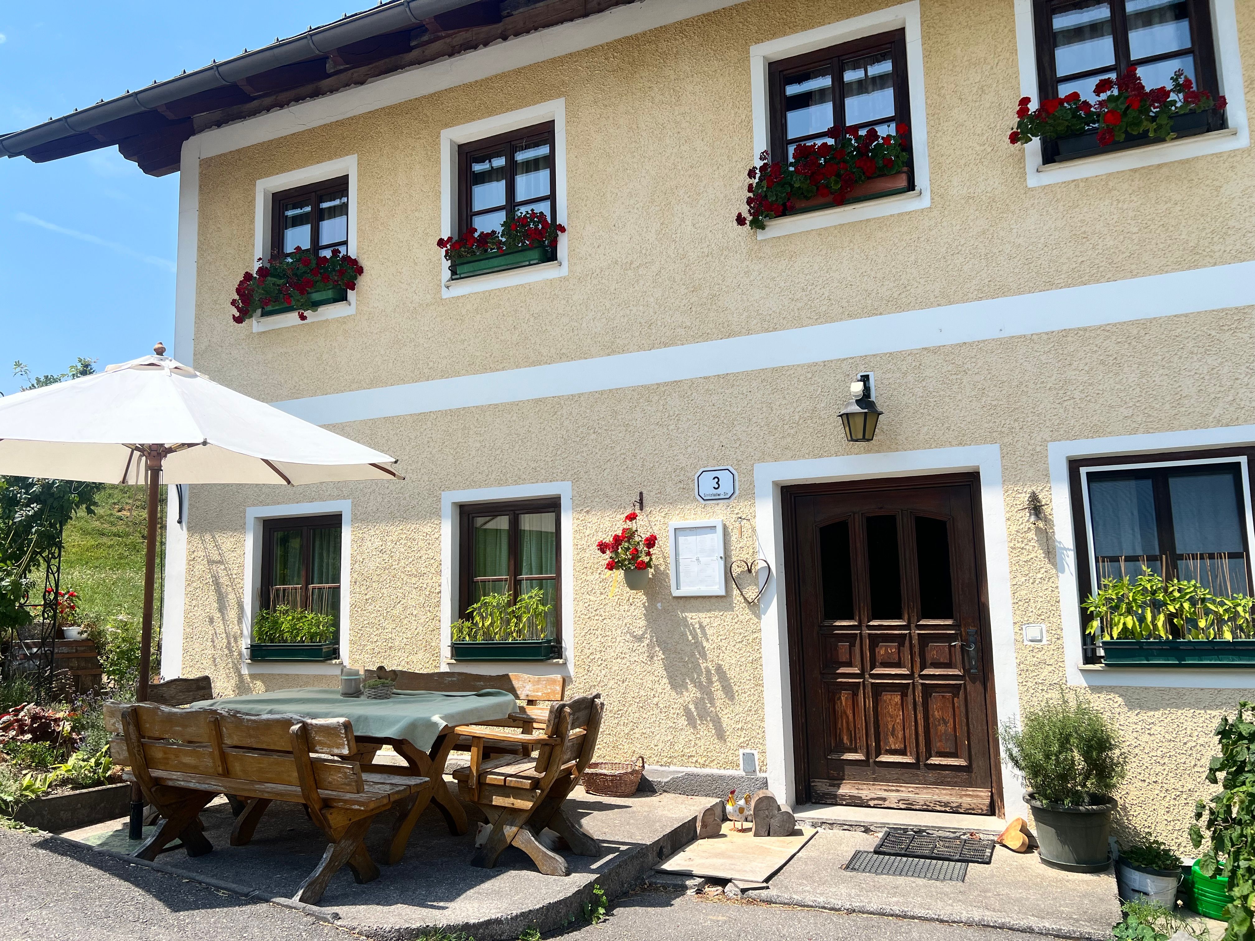 Ein traditionelles Bauernhaus mit gelber Fassade und roten Blumen in den Fenstern. Vor dem Haus steht ein Holztisch mit Bänken und einem Sonnenschirm.
