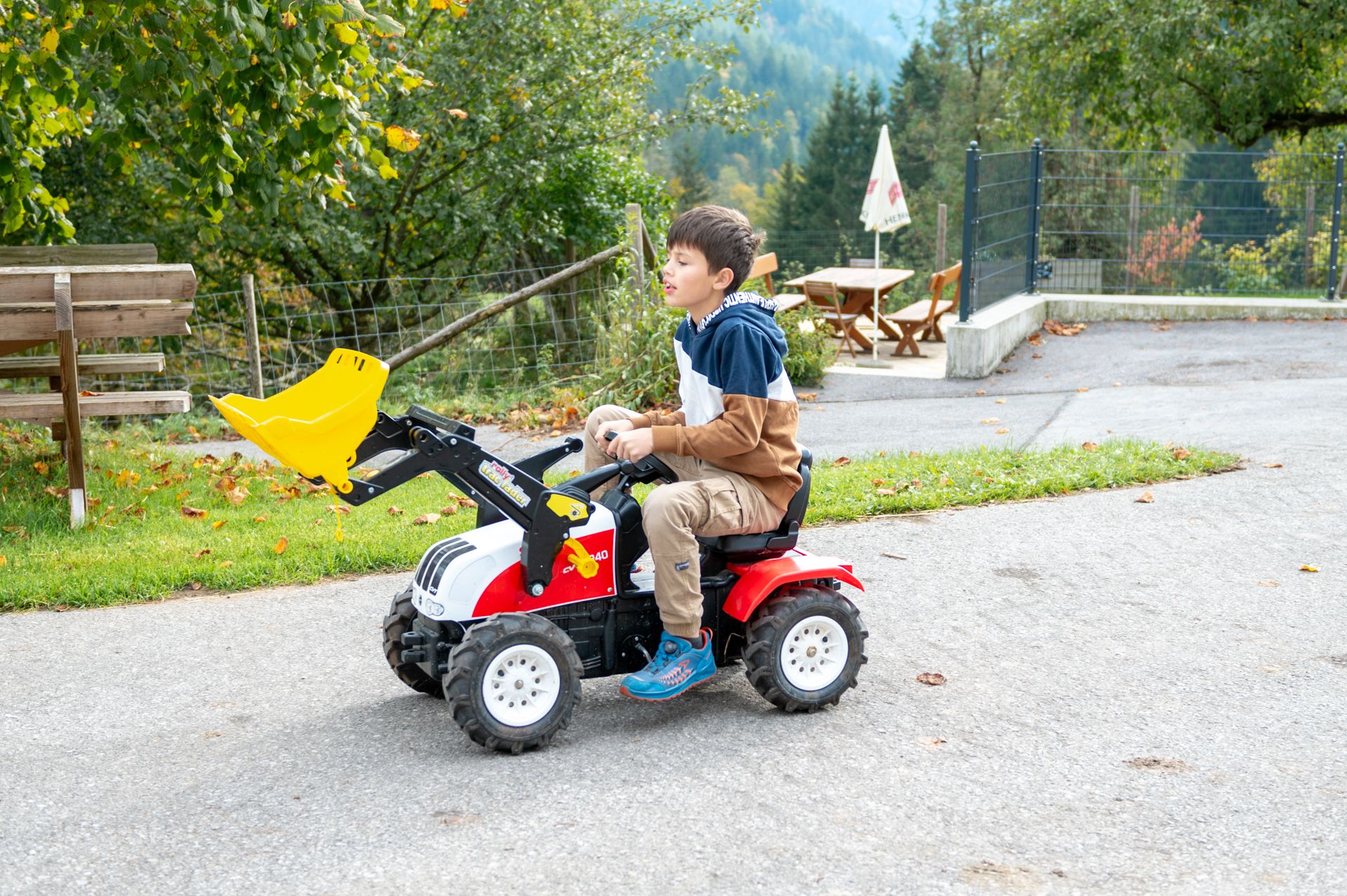 Ein Kind fährt auf einem kleinen Trettraktor mit Schaufel auf einem Weg im Freien.