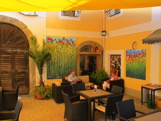 Ein gemütlicher Innenhof eines Cafés mit gelben Wänden, Pflanzen und Gemälden. Zwei Personen sitzen an einem Tisch.
