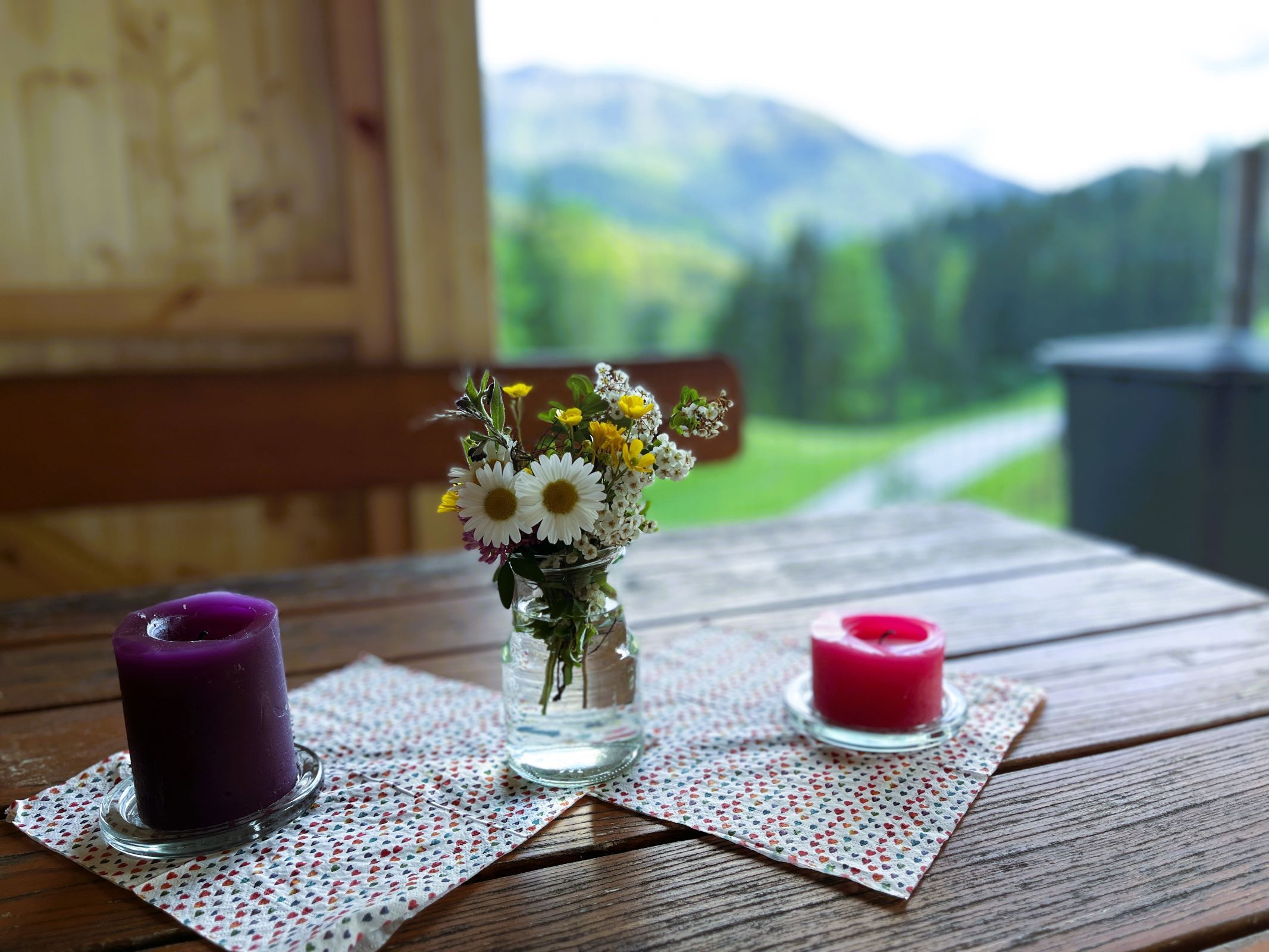 Ein Holztisch mit zwei Kerzen und einem kleinen Blumenstrauß in einem Glasgefäß, im Hintergrund eine verschwommene Berglandschaft.